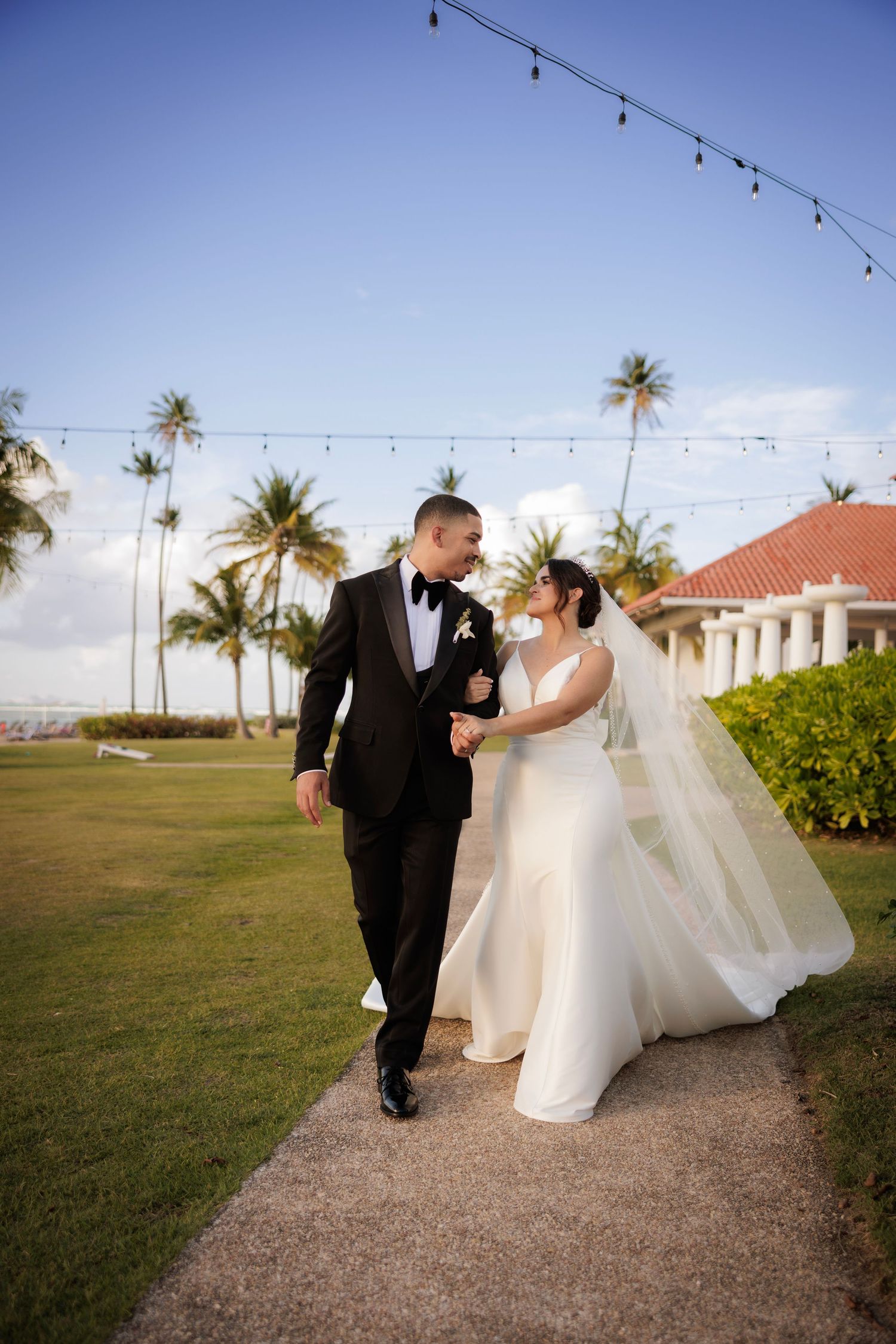 A couple in wedding attire walks along a palm-lined path with string lights overhead and a resort building in the background.