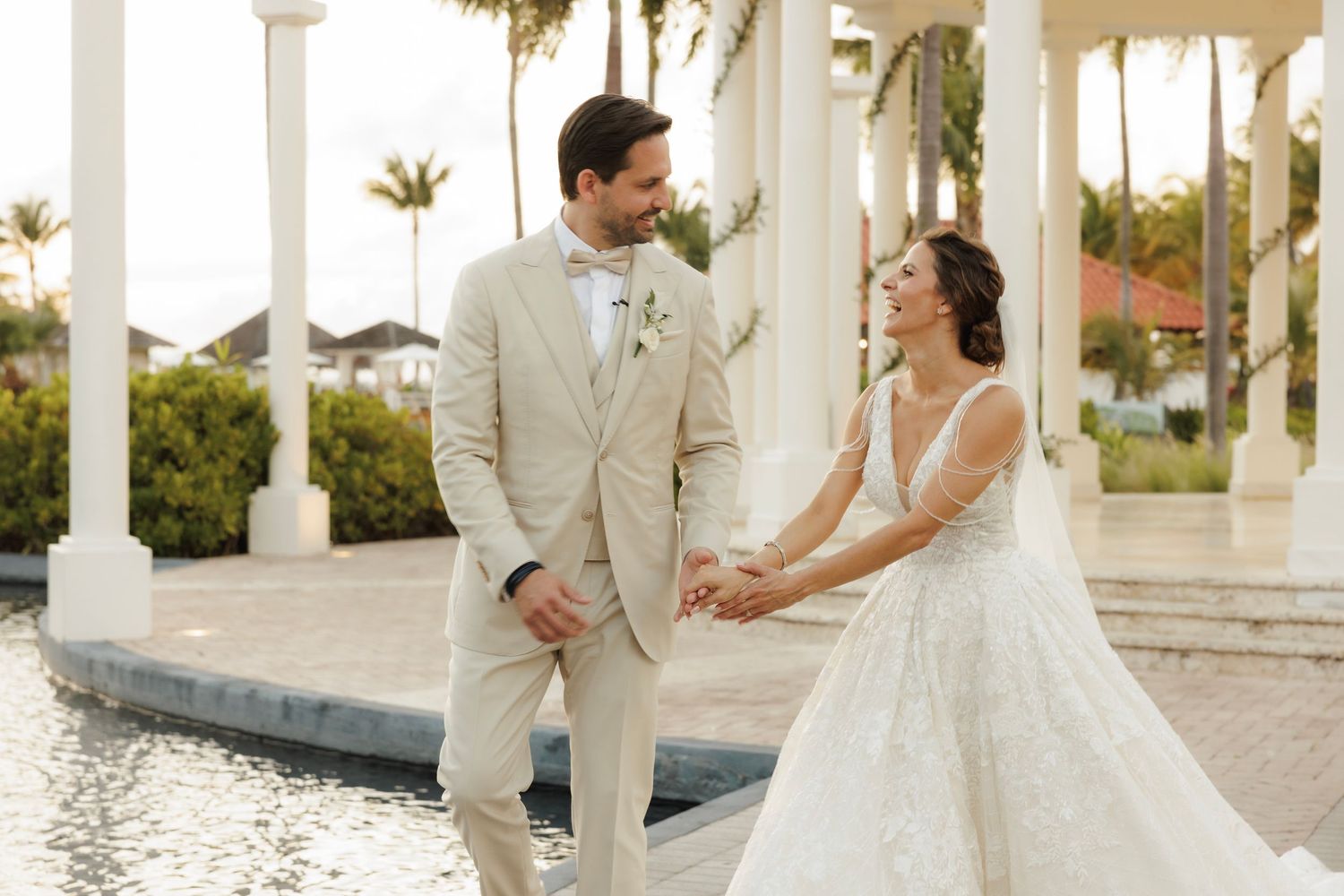 A couple in wedding attire share a joyful moment beside a tropical garden fountain at sunset.