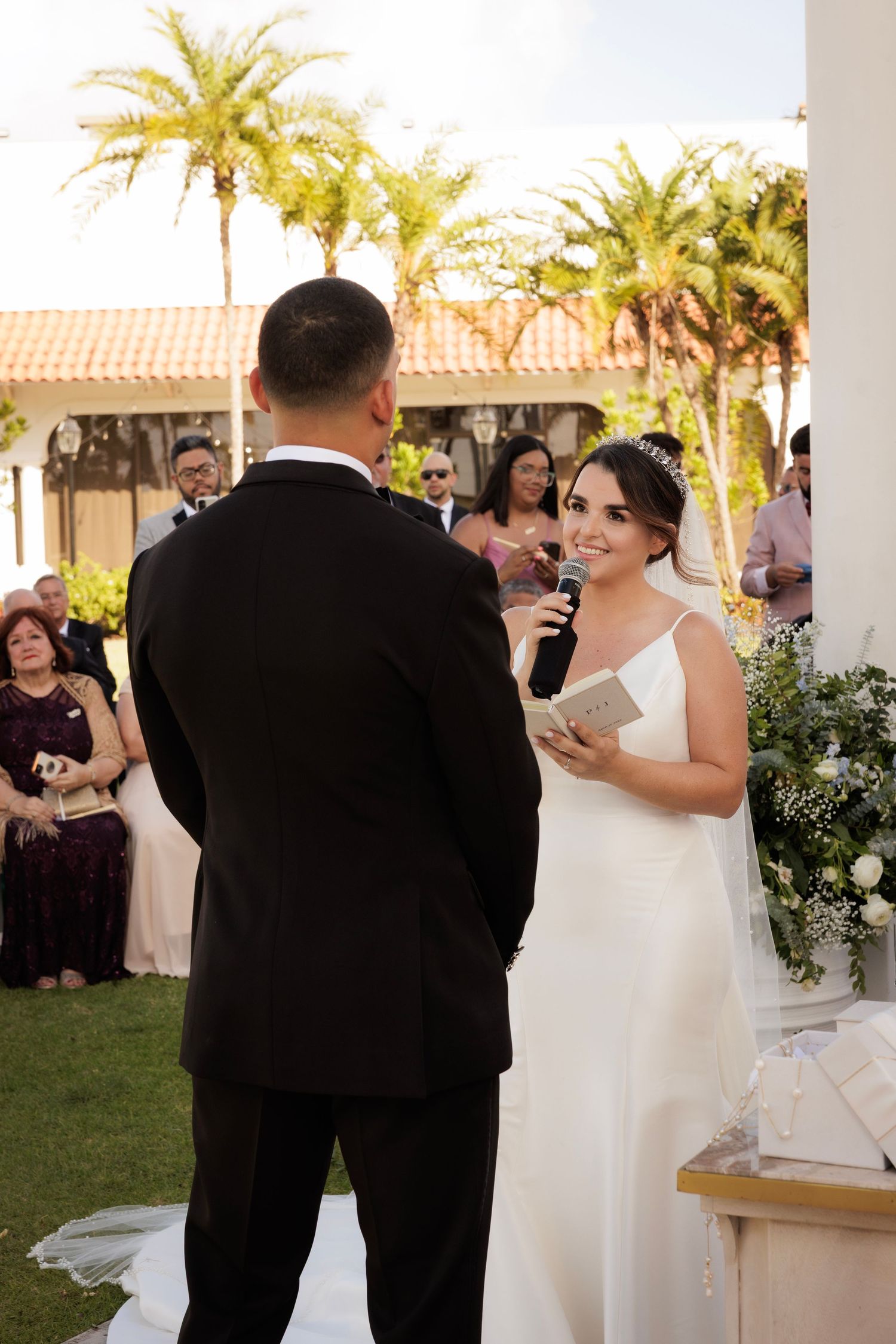 Romantic wedding ceremony under palm trees with a bride in white gown and groom in black tuxedo exchanging vows.