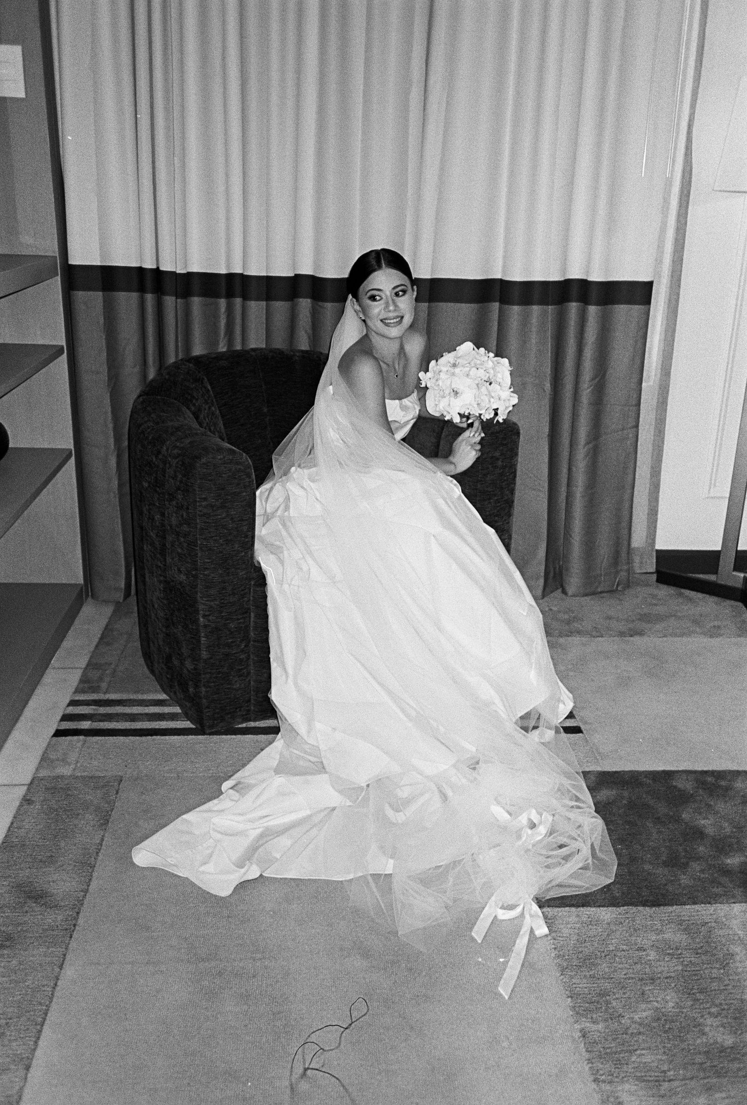 Bride in white ball gown with veil holding bouquet, seated in chair, black and white wedding photo.