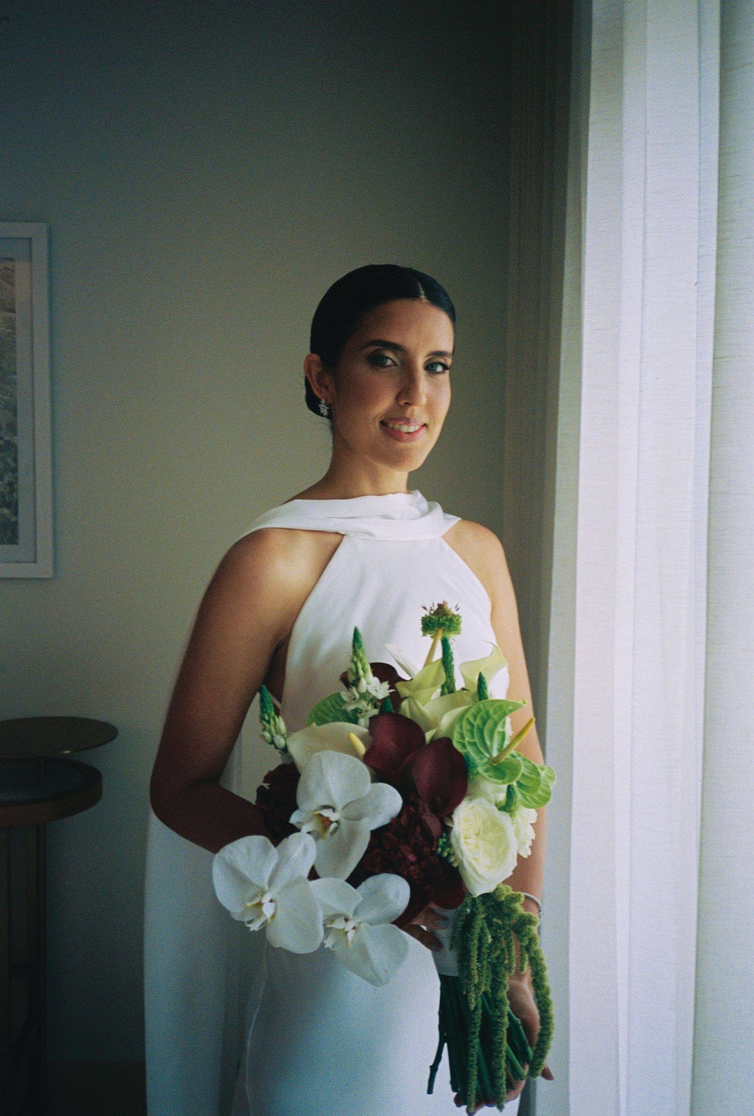 Bride in white halter gown holding bouquet of white orchids and green flowers by a window.