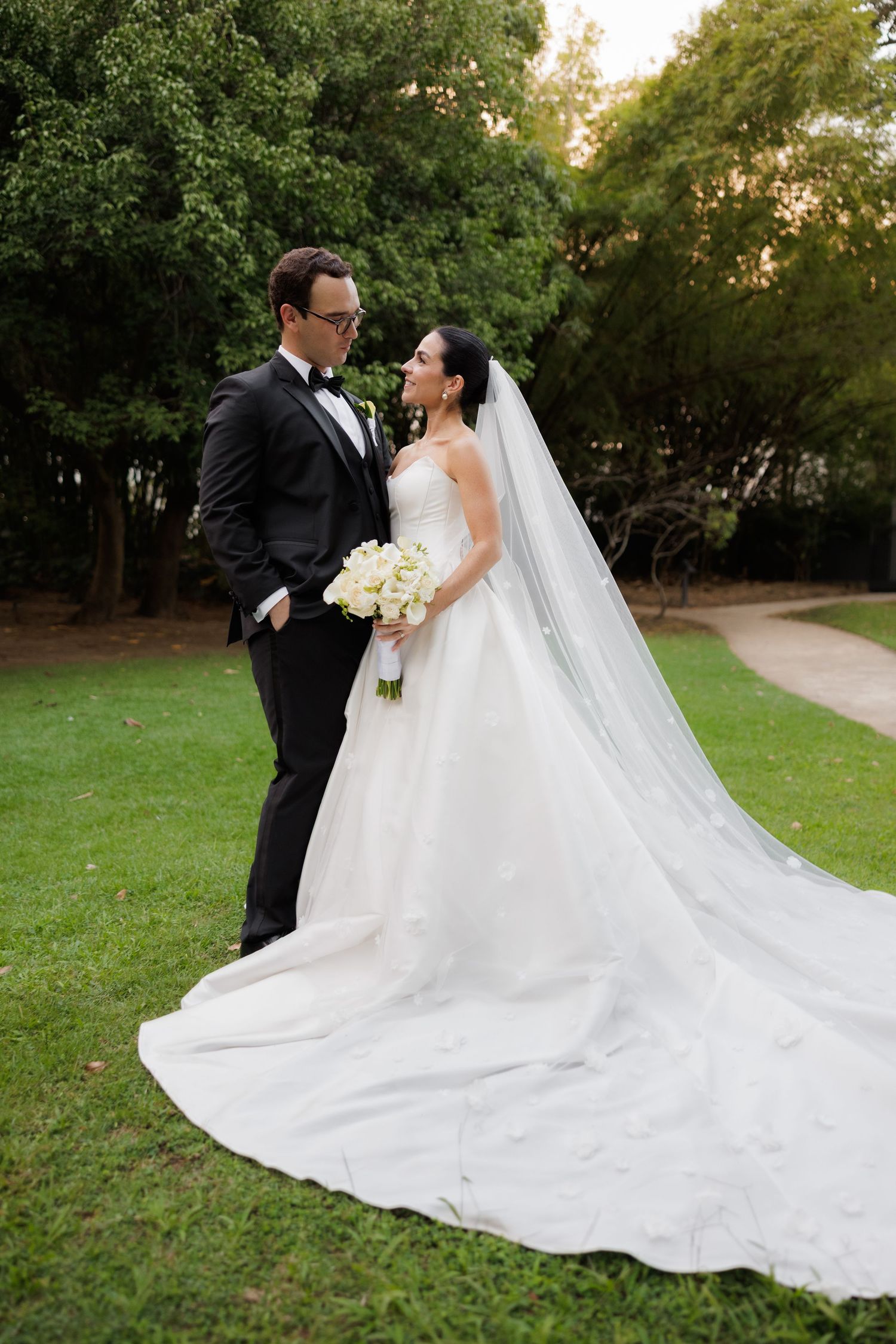 Romantic wedding couple poses in lush garden setting with bride's flowing white gown creating elegant silhouettes at sunset.