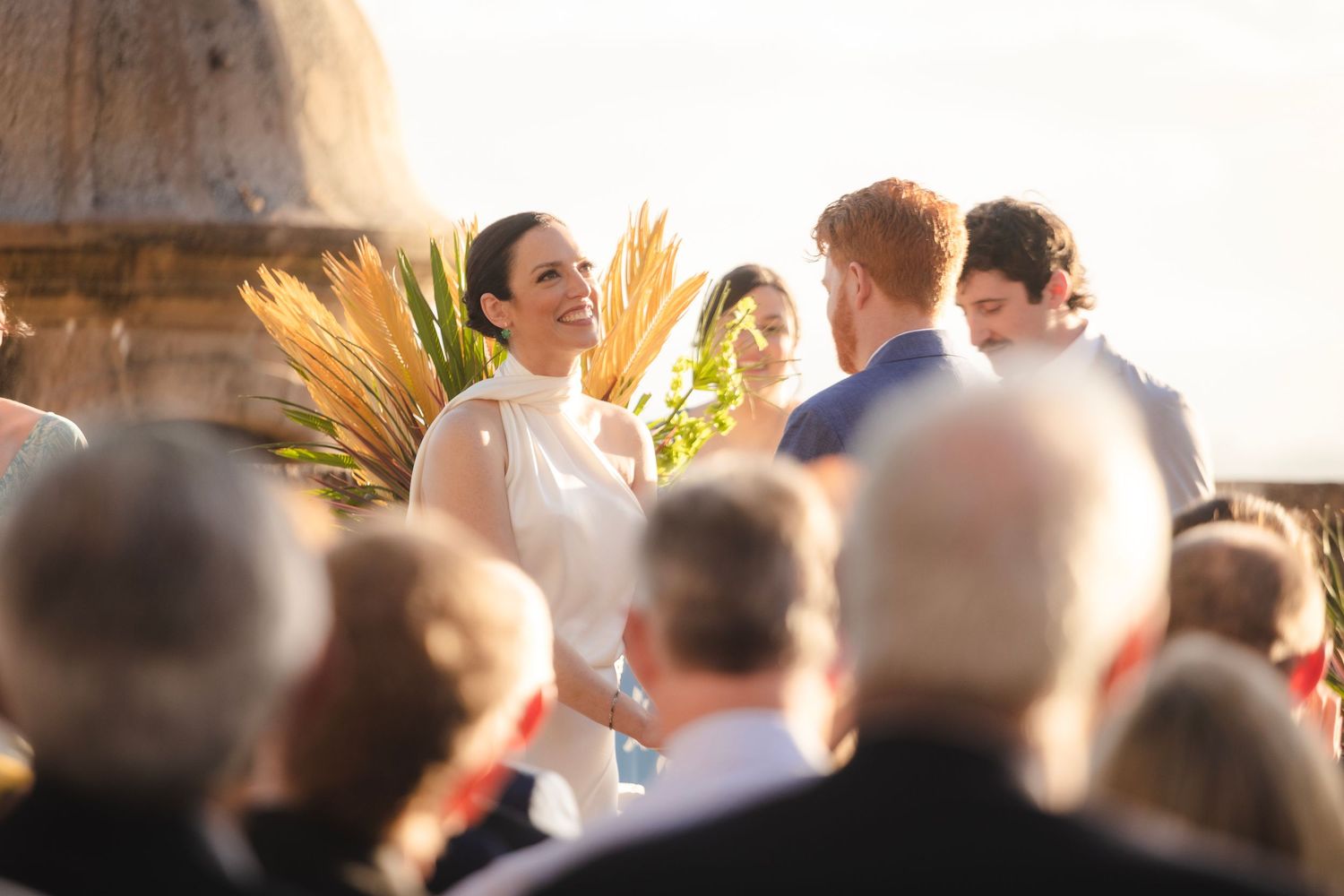 Wedding guests gather outdoors during golden hour creating silhouettes against the bright sky.
