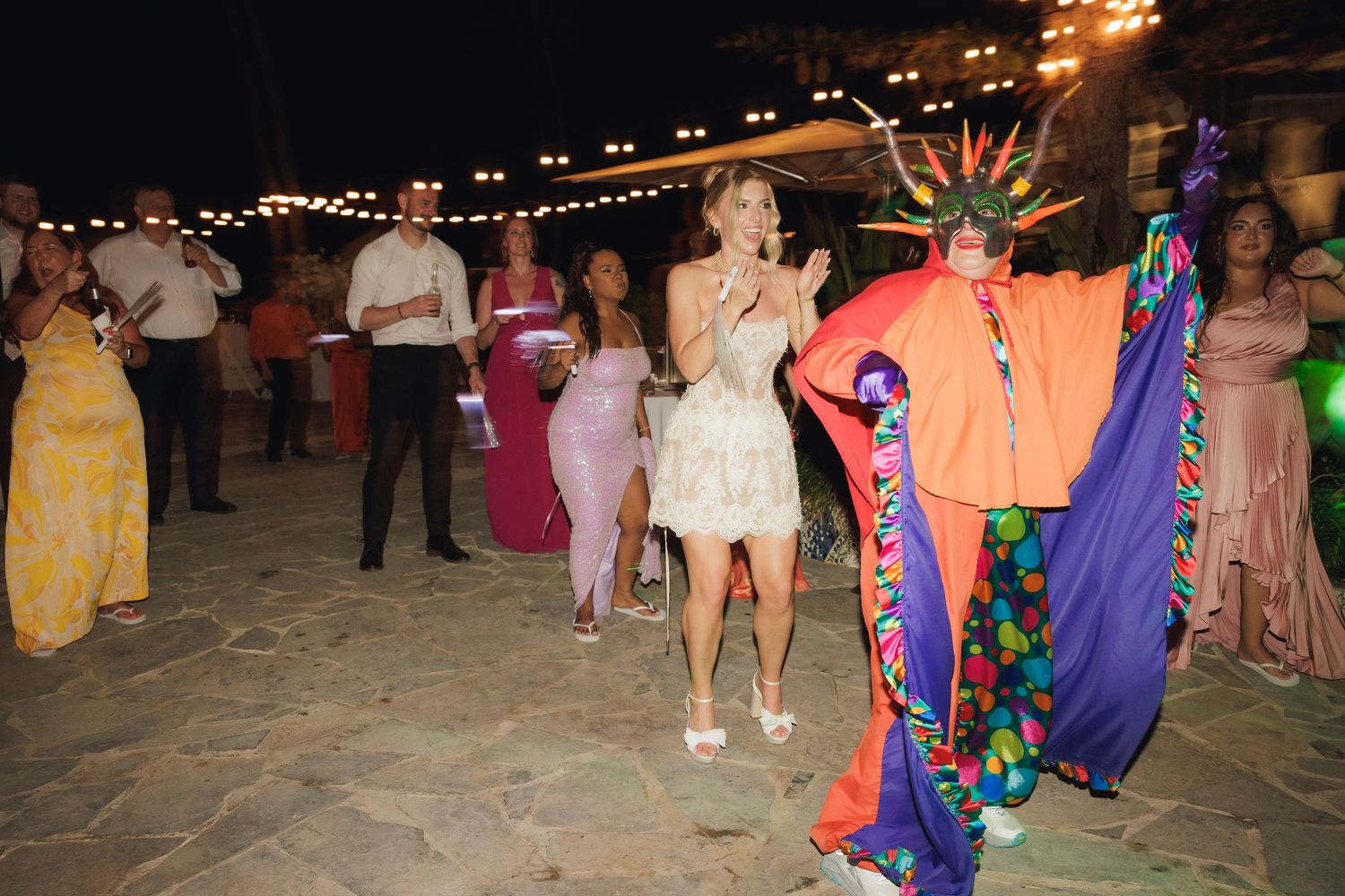 Party guests dancing on a stone patio at night with overhead string lights and colorful party decorations.