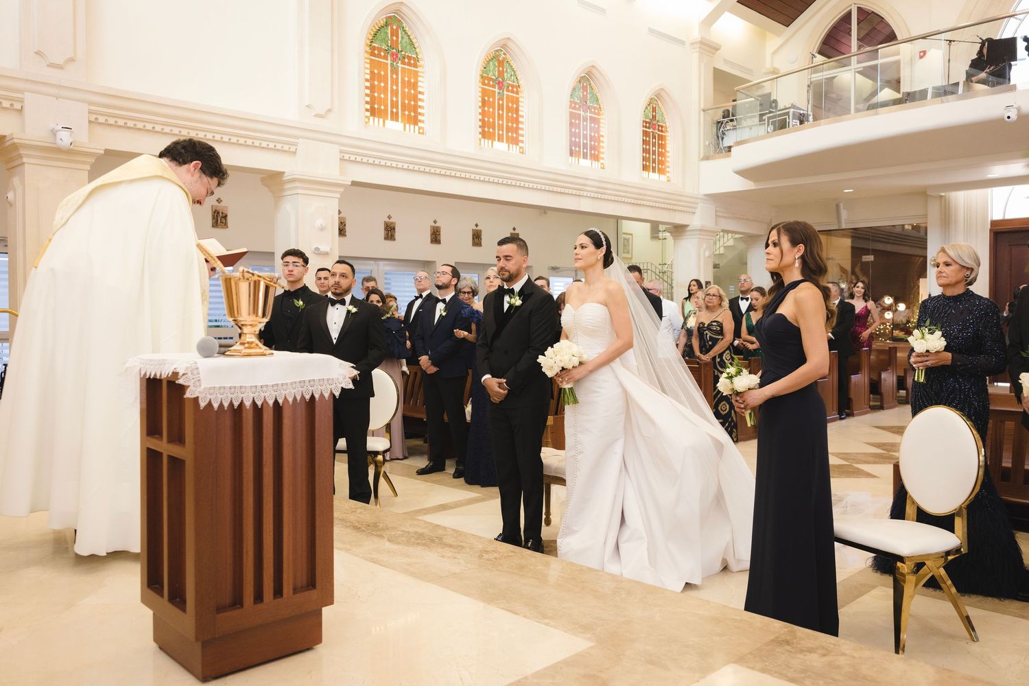 Catholic wedding ceremony taking place inside a bright white church with stained glass windows and traditional altar setup.