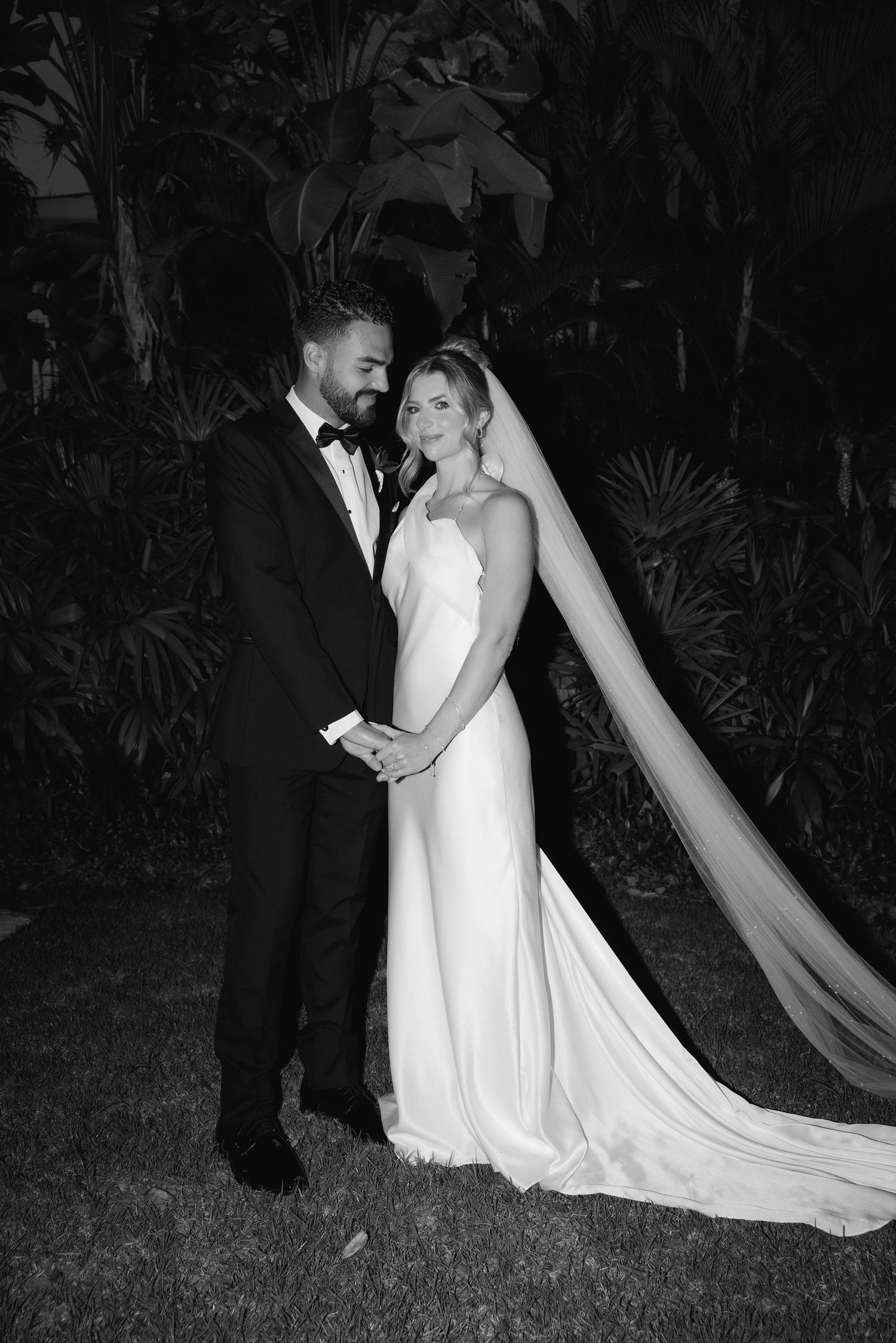 A romantic black and white photo series of a bride and groom embracing outdoors at night with a flowing wedding dress train.
