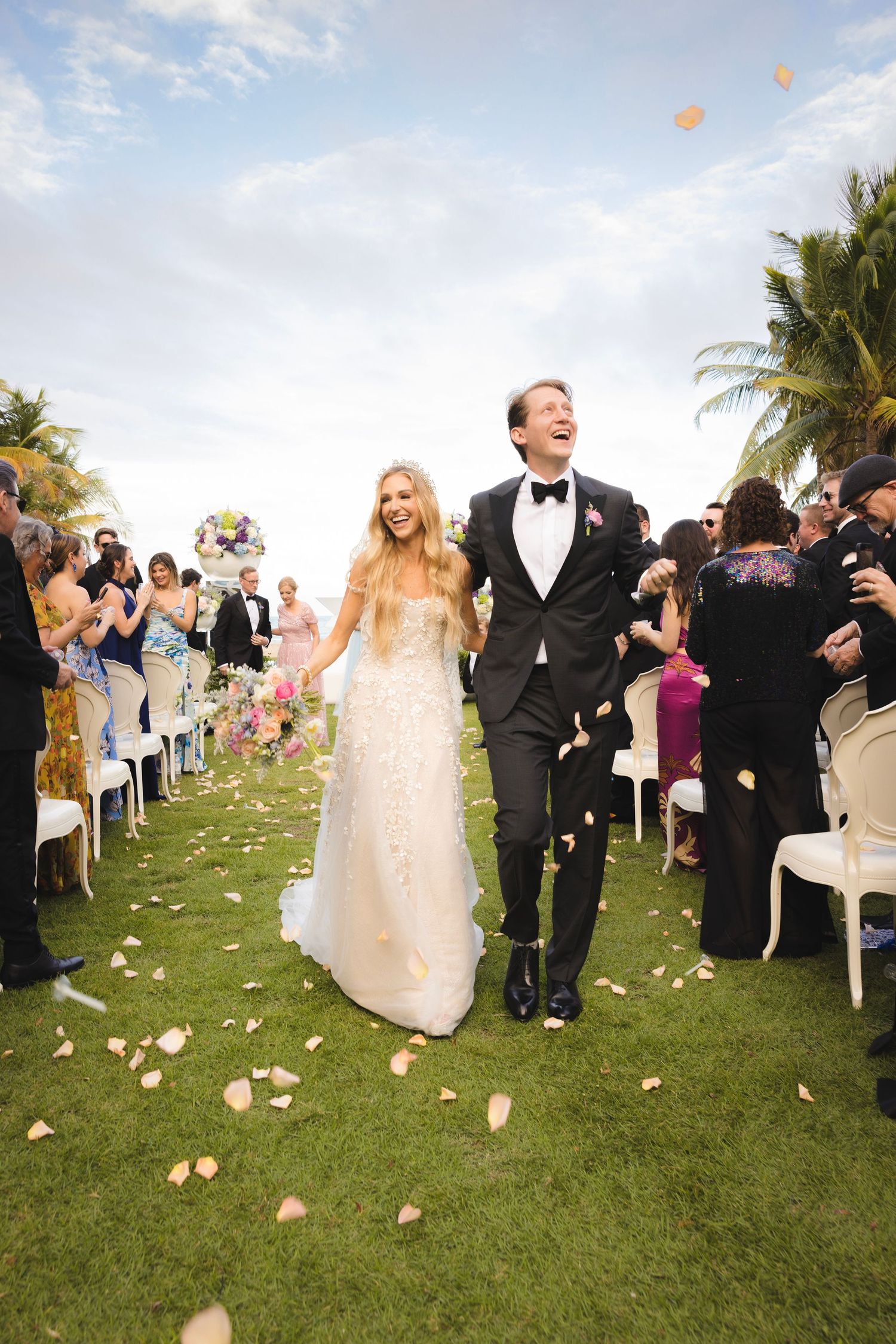 Newlyweds celebrate while walking down aisle at sunny outdoor beach wedding ceremony with palm trees and flower petals.