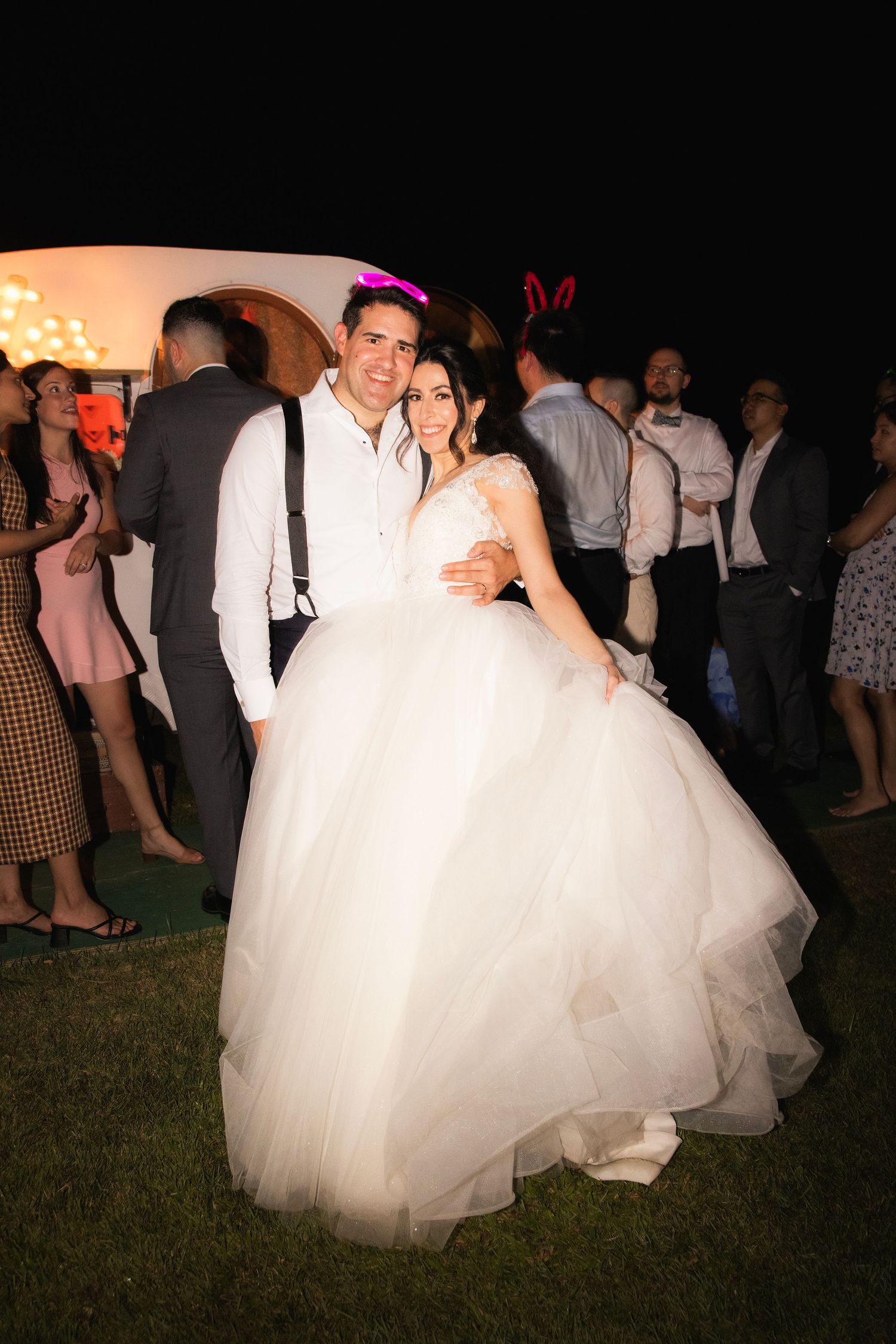 Wedding guests dancing and celebrating with bride in white gown under dimly lit outdoor evening setting.