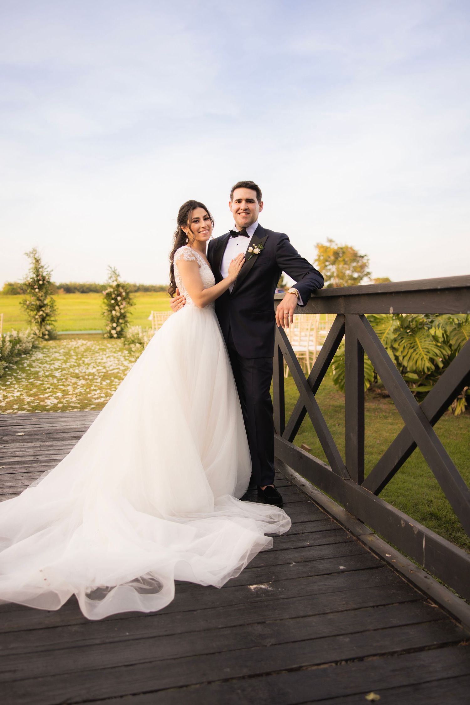 Couple in wedding attire poses together on a wooden bridge deck with scenic countryside views.