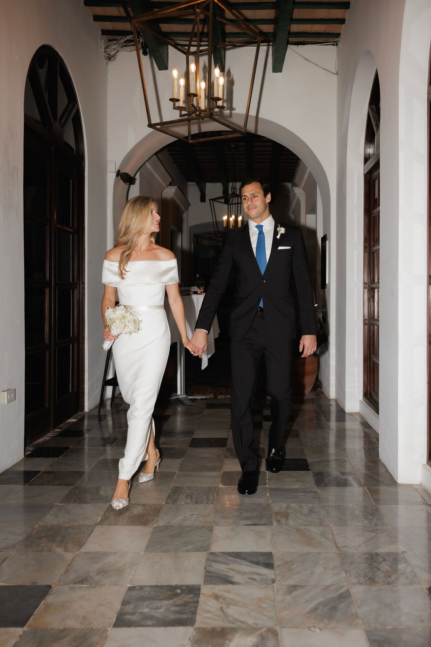 A couple in formal attire walks down an elegant marble hallway with arched walls and decorative lighting.