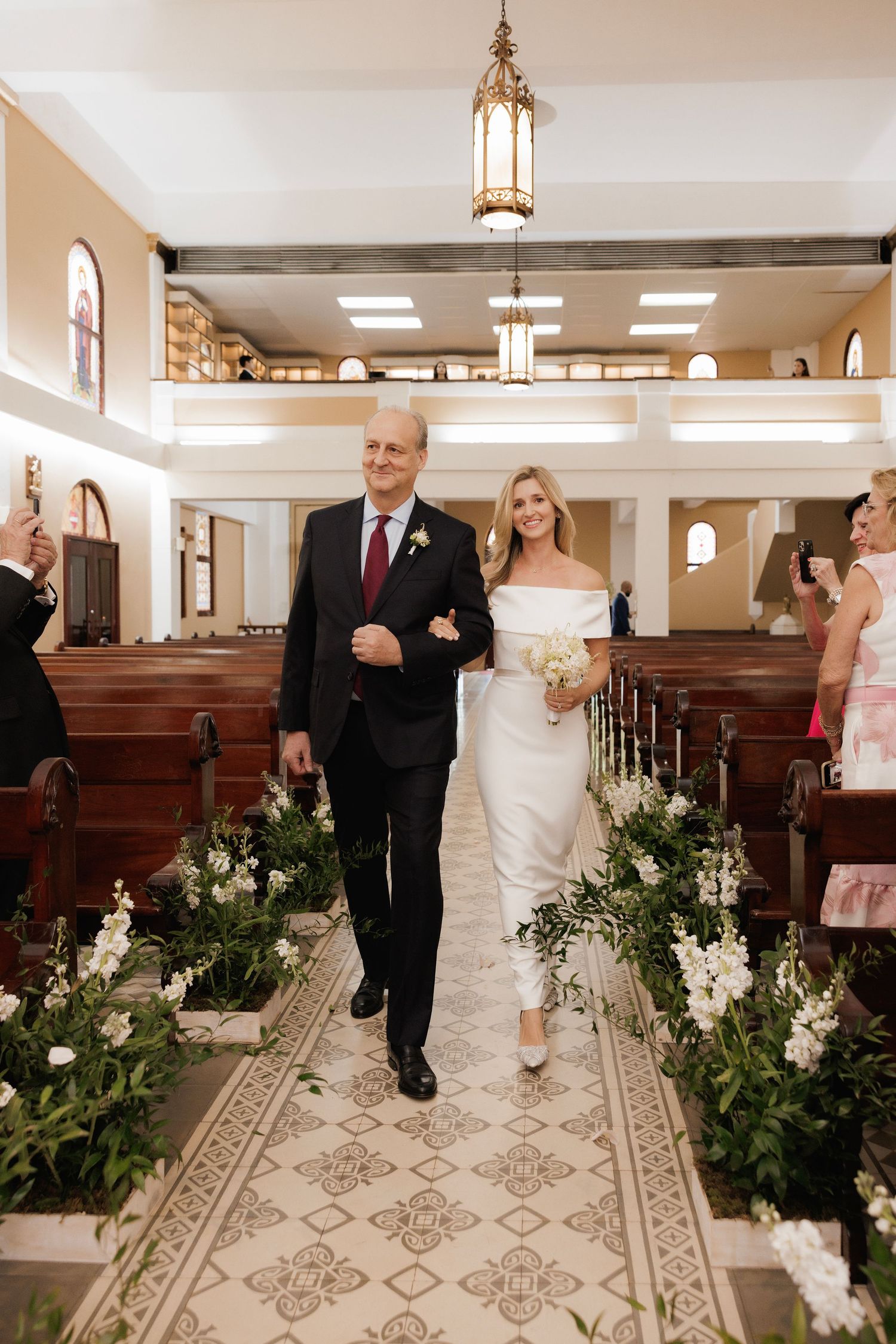 A wedding procession walks down a carpeted church aisle decorated with elegant white floral arrangements and lantern lighting.