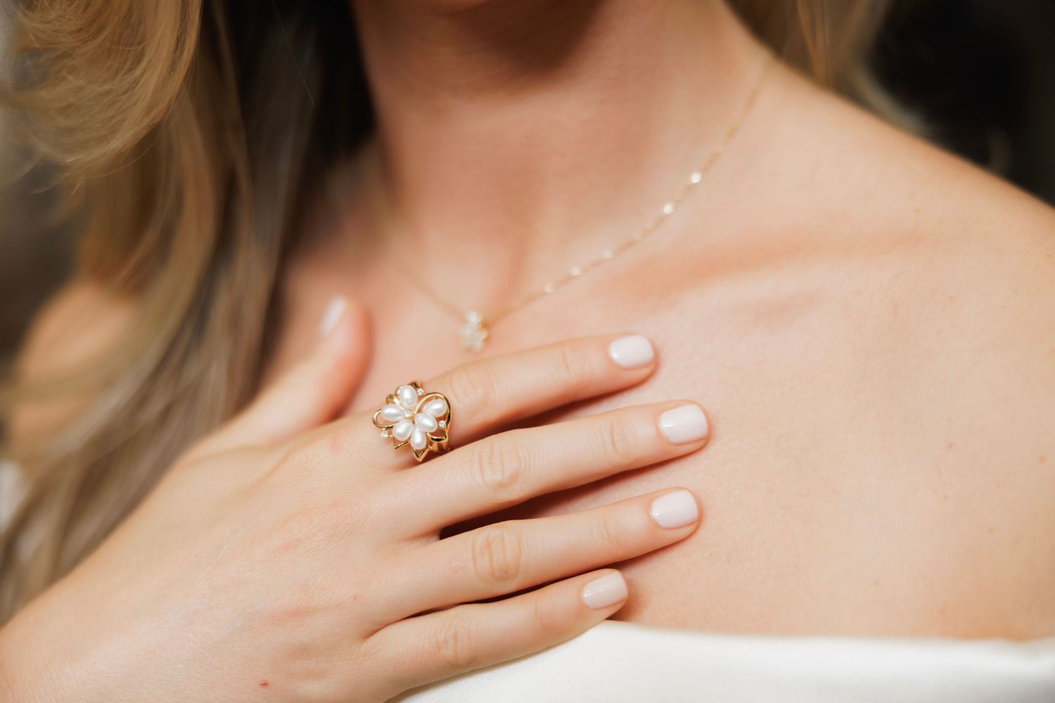 Elegant hand wearing a shimmering floral ring with pale pink nail polish and a delicate necklace against soft white fabric.