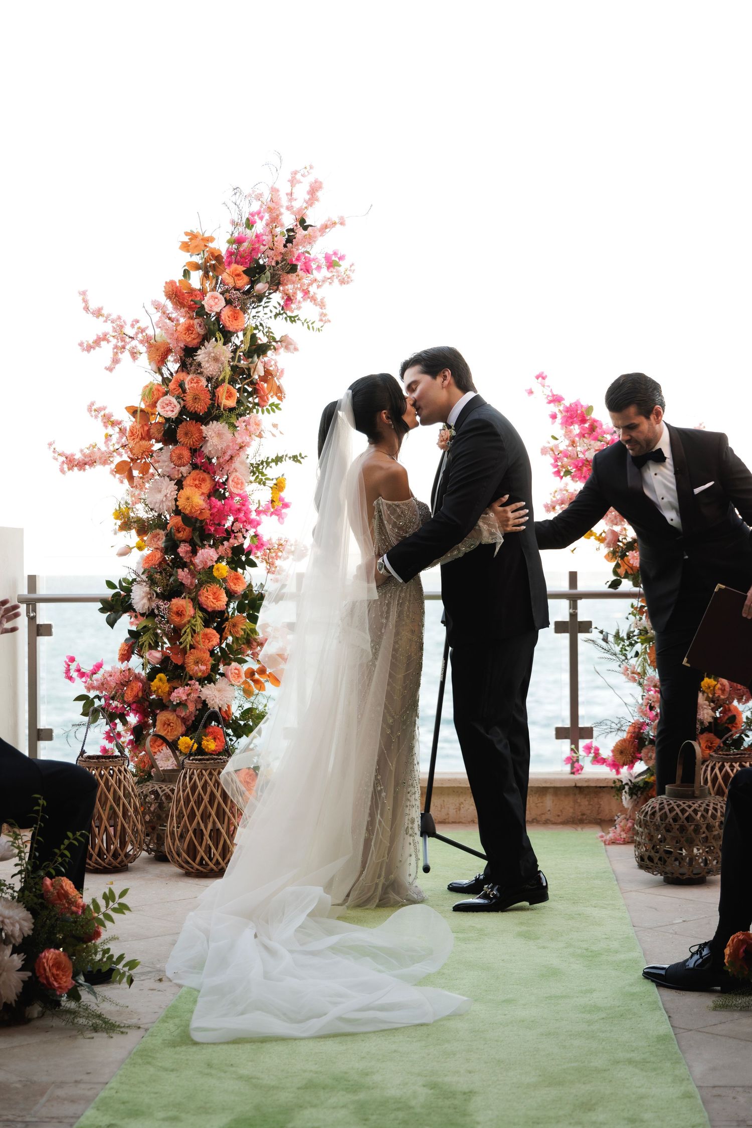 A wedding ceremony on a green-carpeted terrace with stunning coral and pink floral arrangements framing the romantic moment.