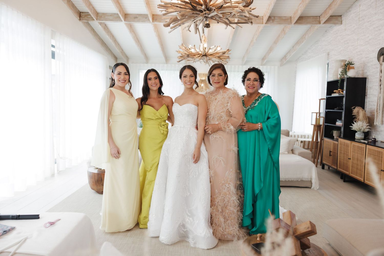 A group in yellow, white, beige and turquoise dresses laugh together in an elegant room with exposed beams.