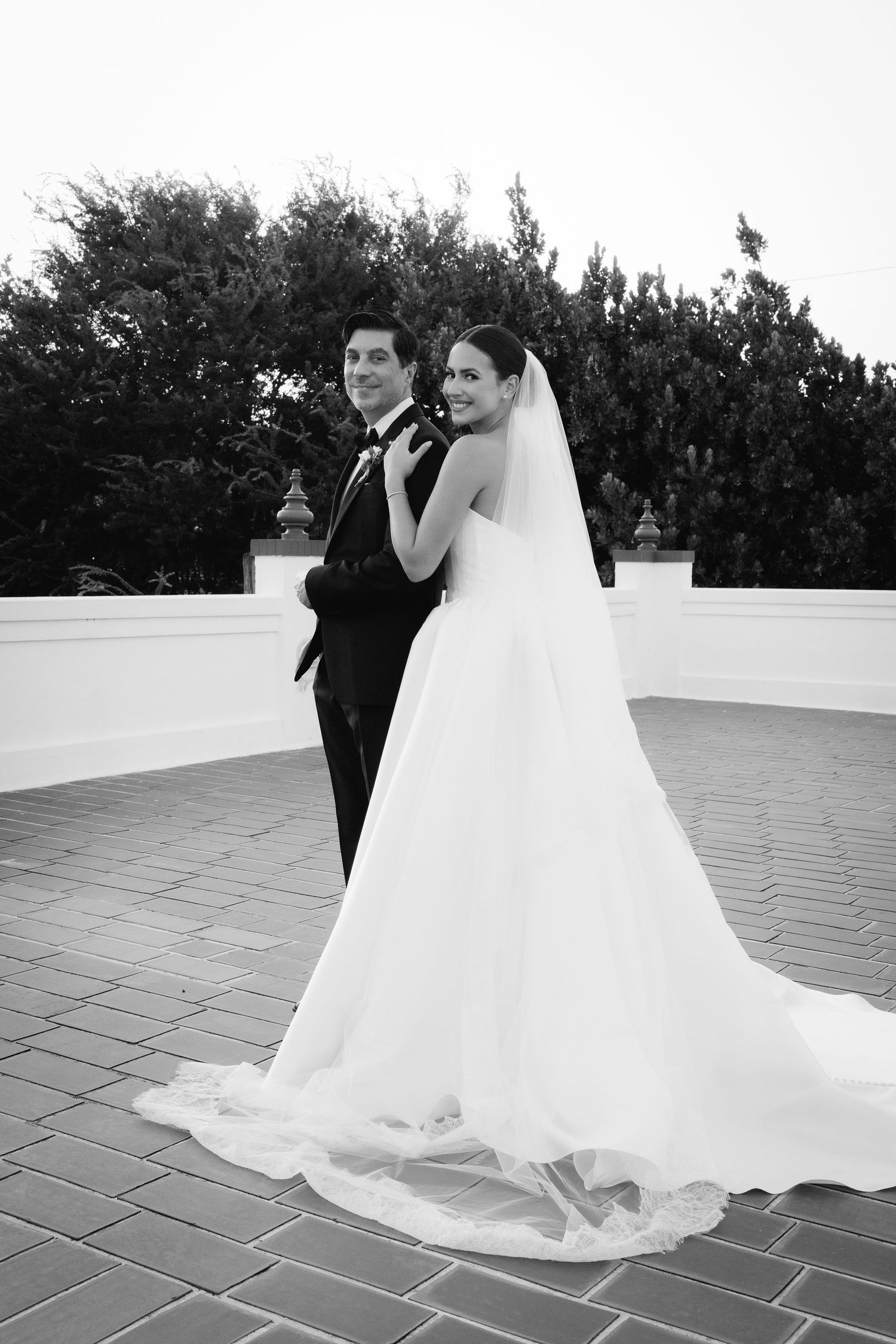 A bride in a flowing white gown and groom in black tuxedo dance together on a stone terrace.