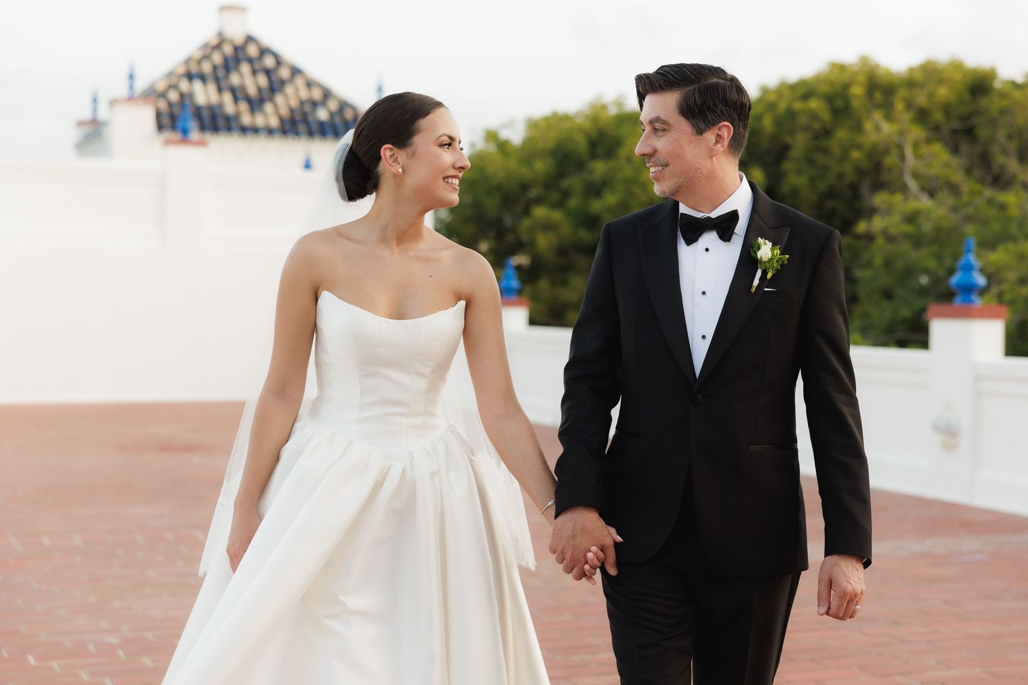 A couple in wedding attire holds hands while walking on a rooftop terrace with white architecture and trees in background.