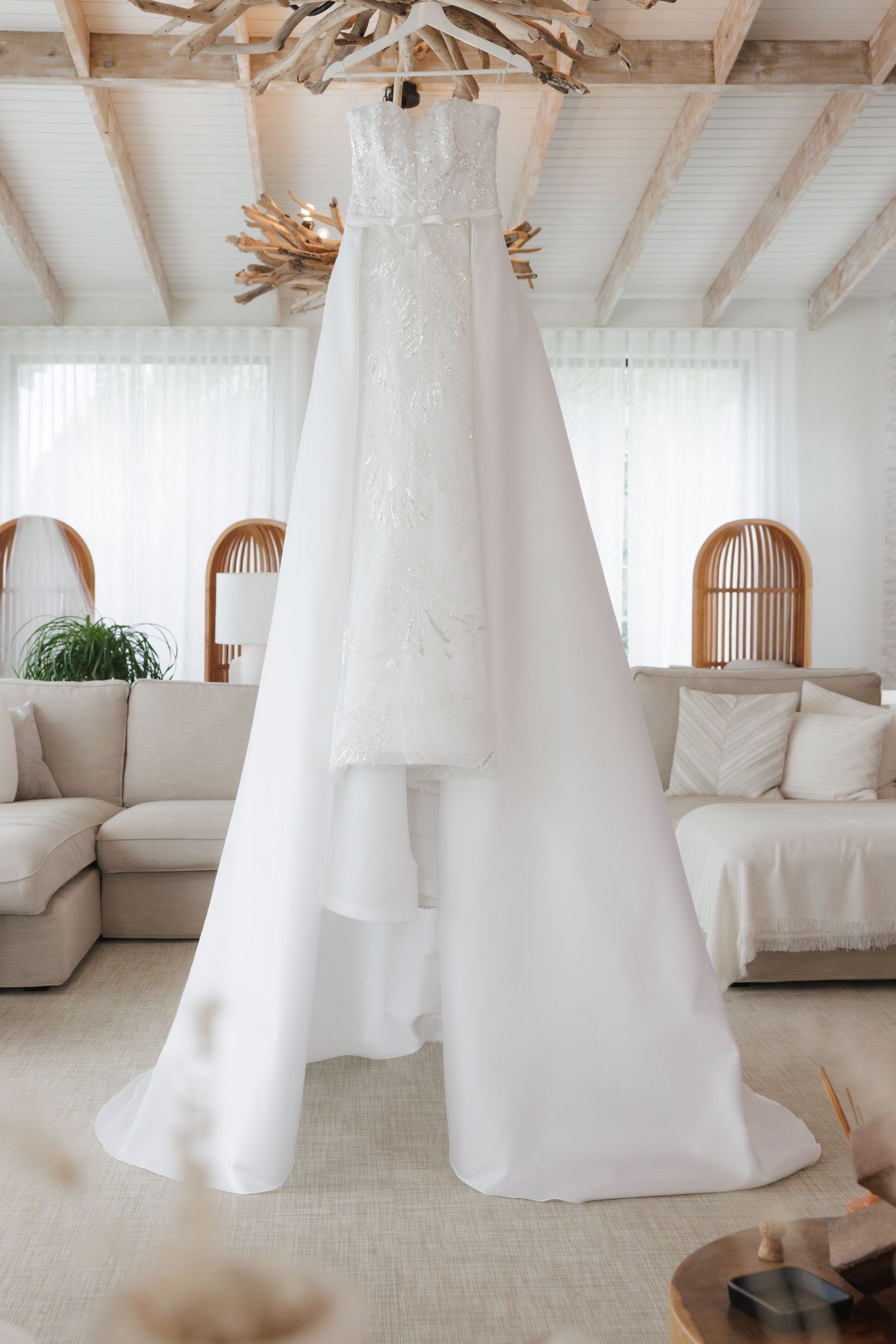An elegant white wedding gown hangs from a rustic wooden beam in a light-filled living room.