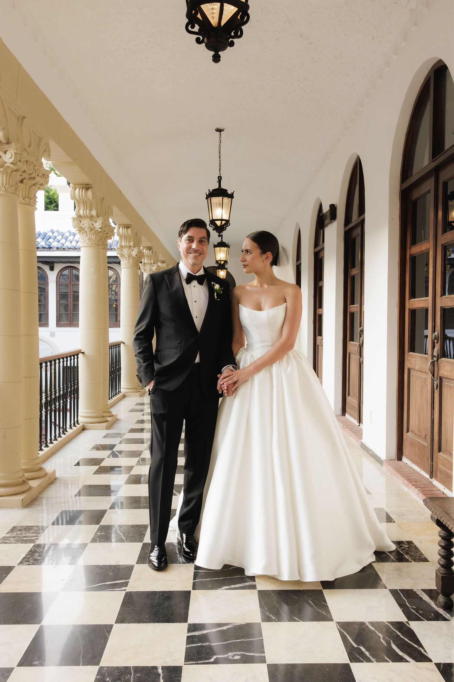 Bride and groom share joyful moments while walking through a columned hallway with checkerboard flooring.