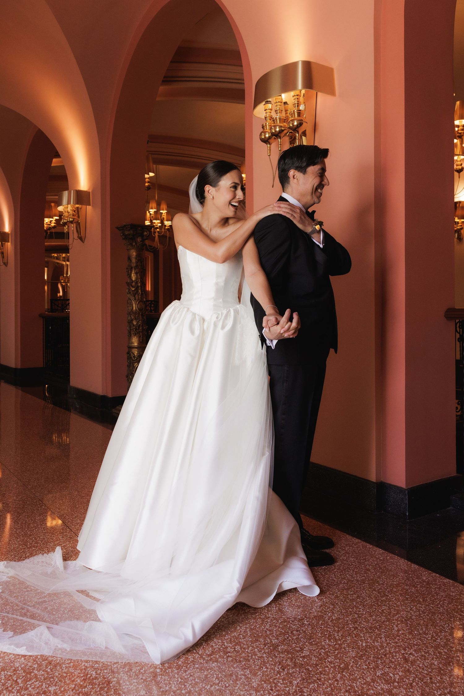 A bride in a flowing white gown and groom in black tuxedo pose against a pink-lit hallway with ornate decor.