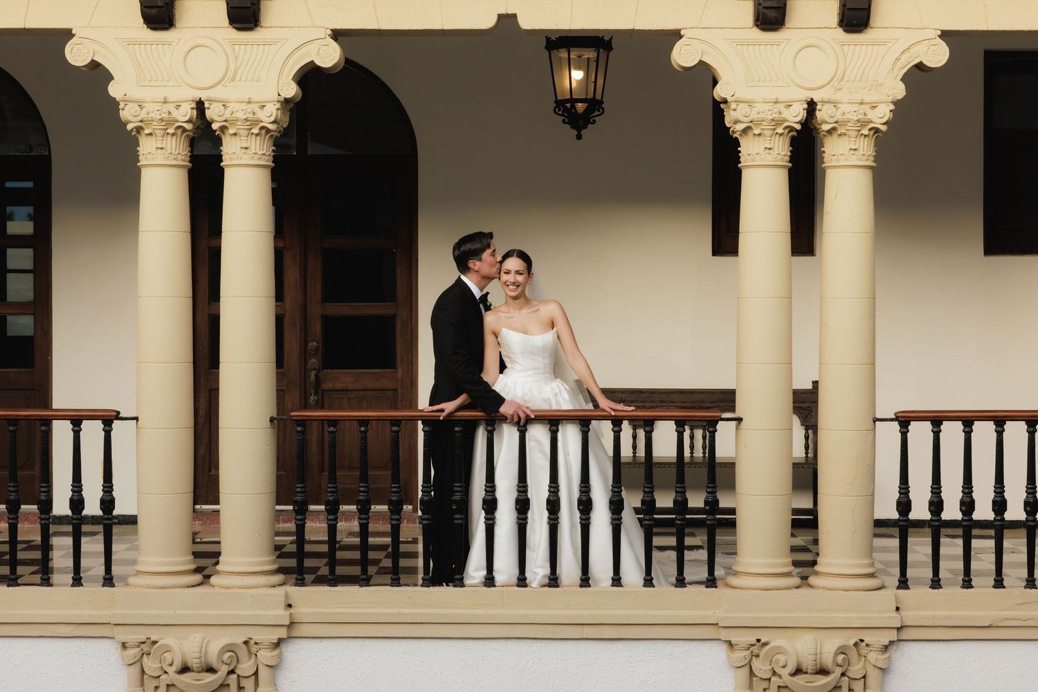Black and white photo of newlyweds strolling along a covered walkway with ornate columns.