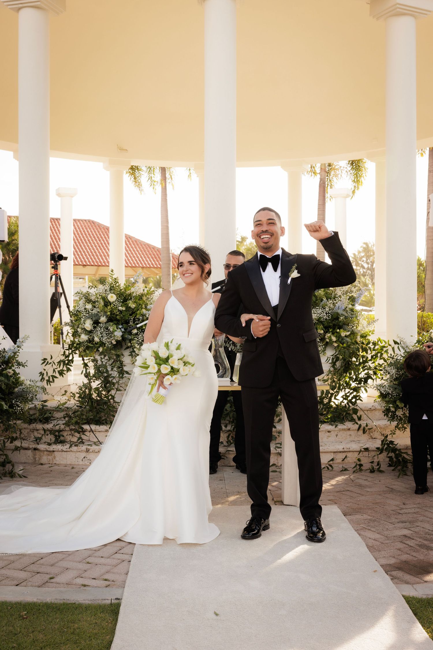 Wedding couple and officiant share joyful moments under elegant white columns during outdoor ceremony.