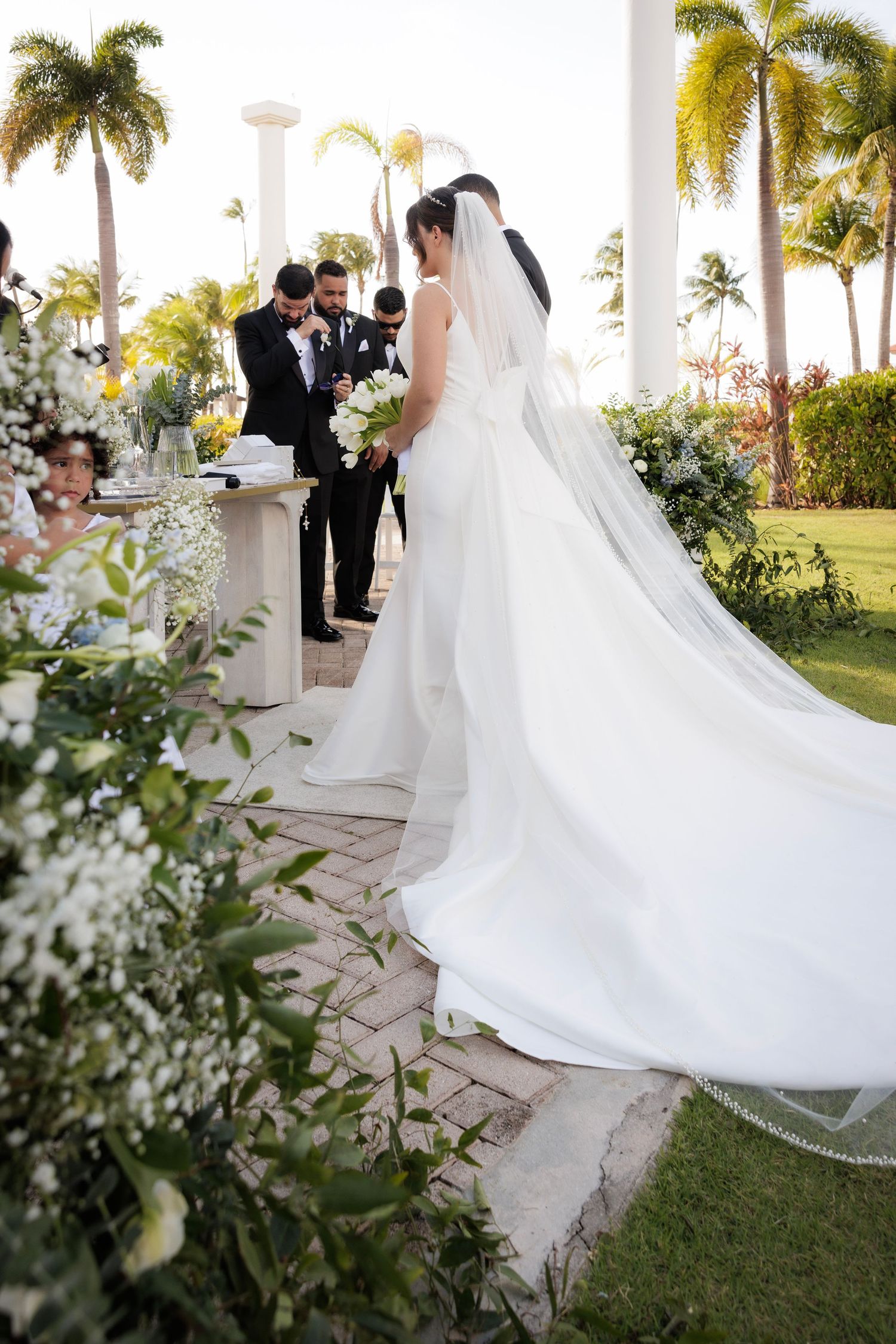 A beautiful outdoor wedding ceremony featuring a bride in an elegant white gown with a flowing train amid tropical palm trees.