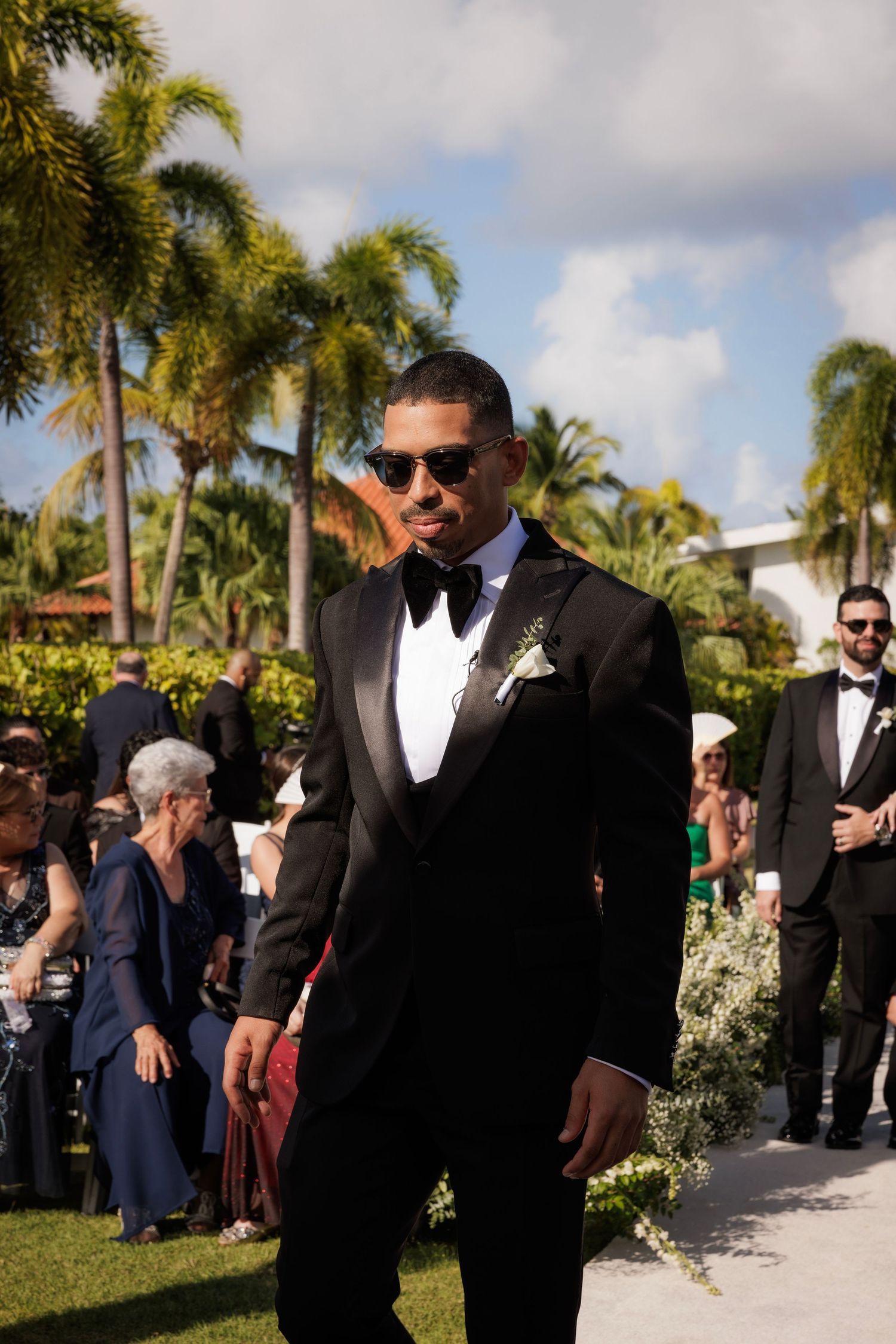 Wedding guests in formal black tuxedos exchange greetings in an elegant outdoor venue lined with palm trees.