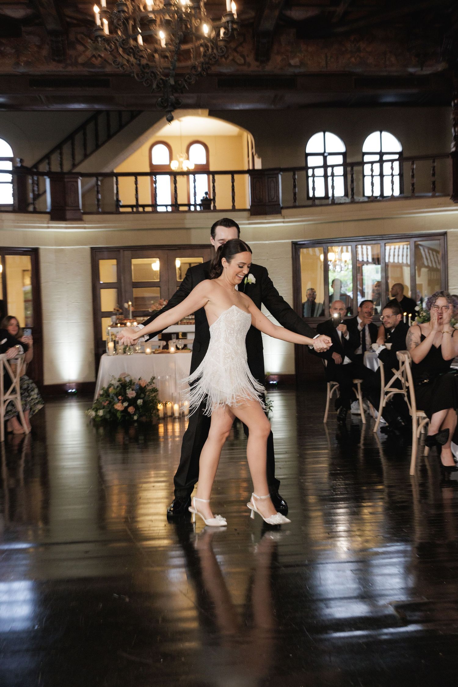 Dancers execute graceful moves on a shiny dance floor while guests watch from the sides of an elegant ballroom.