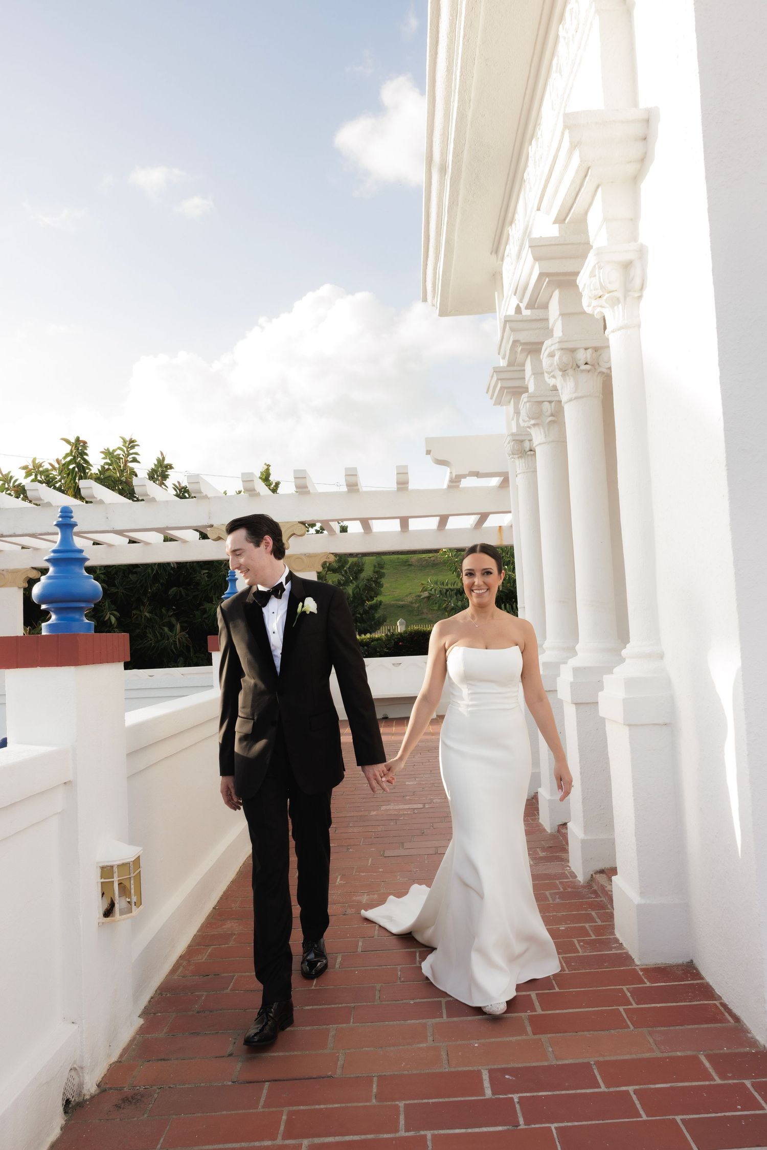 Newlyweds walking along a covered colonnade with white pillars and terracotta flooring.