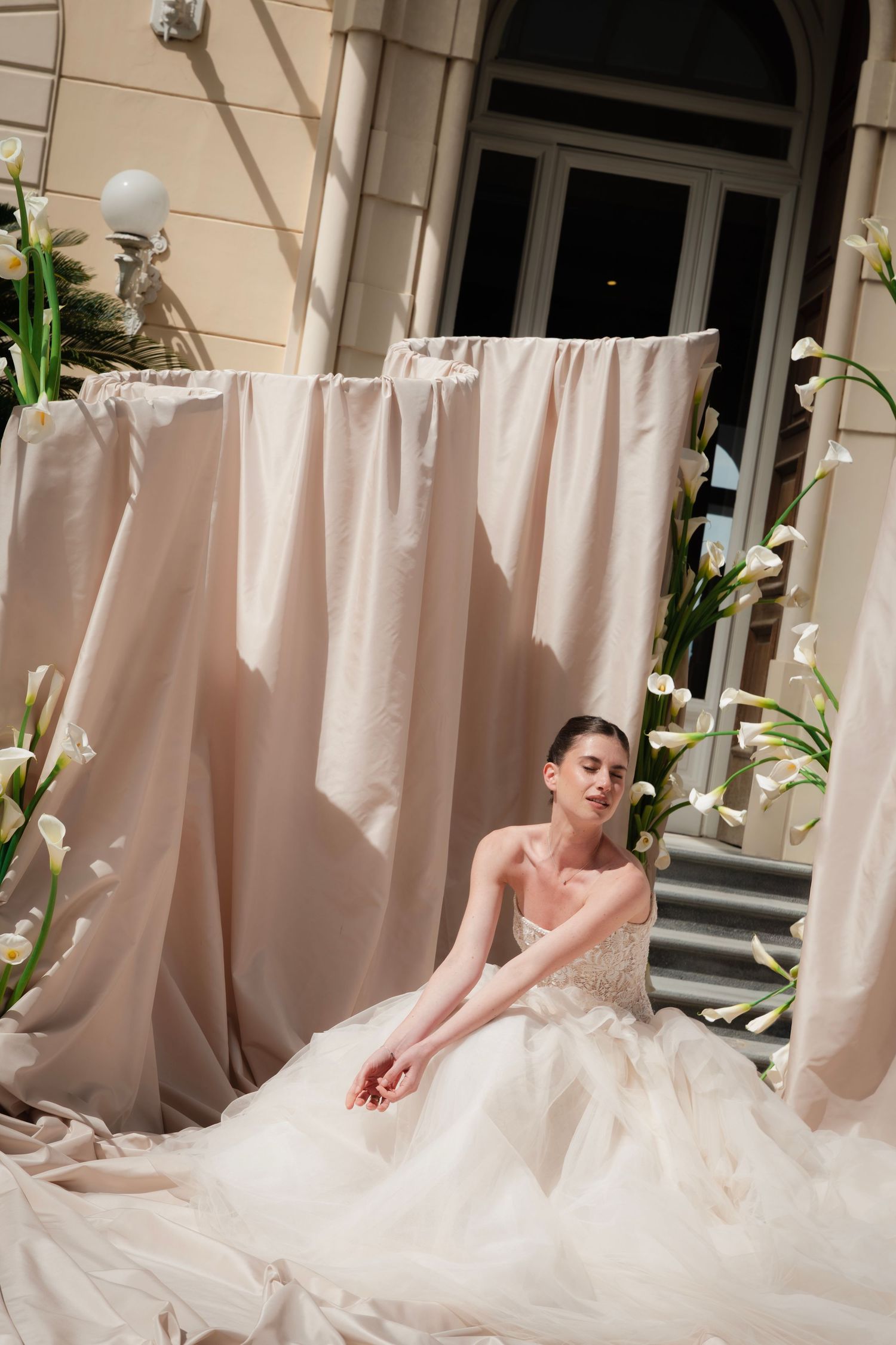 Bride in a flowing tulle wedding gown seated before draped ivory curtains and white calla lilies outdoors.