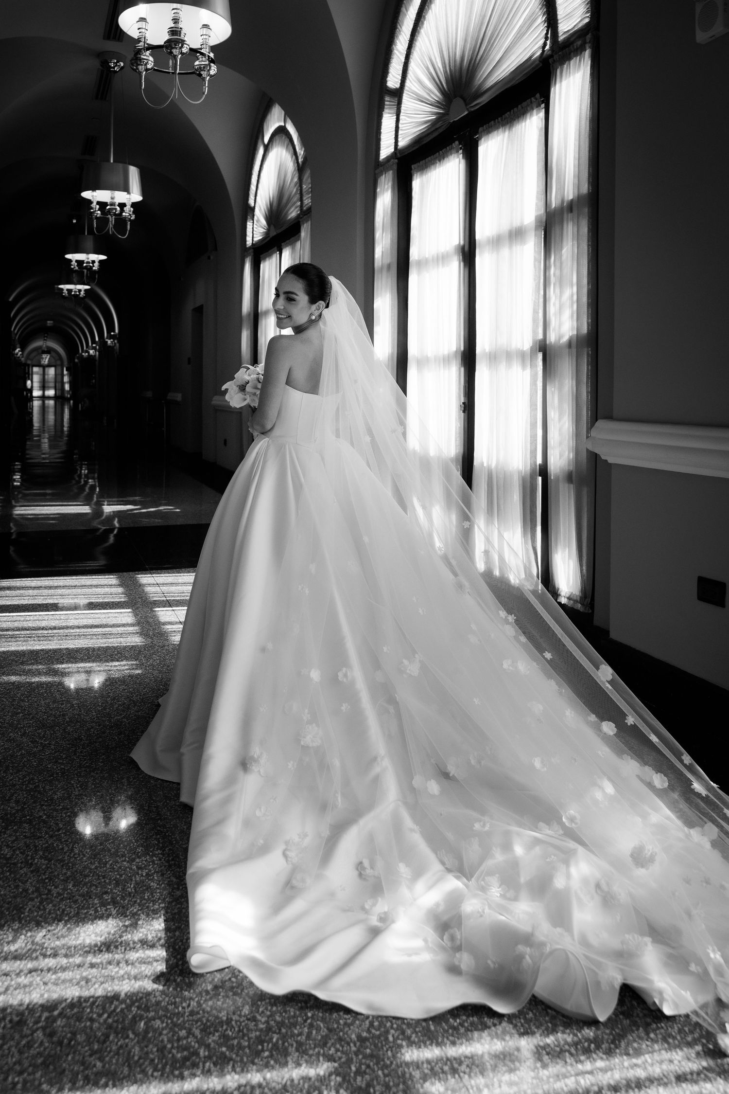 Black and white photo of a bride in a flowing ball gown with veil, standing by sunlit windows in an elegant hall.