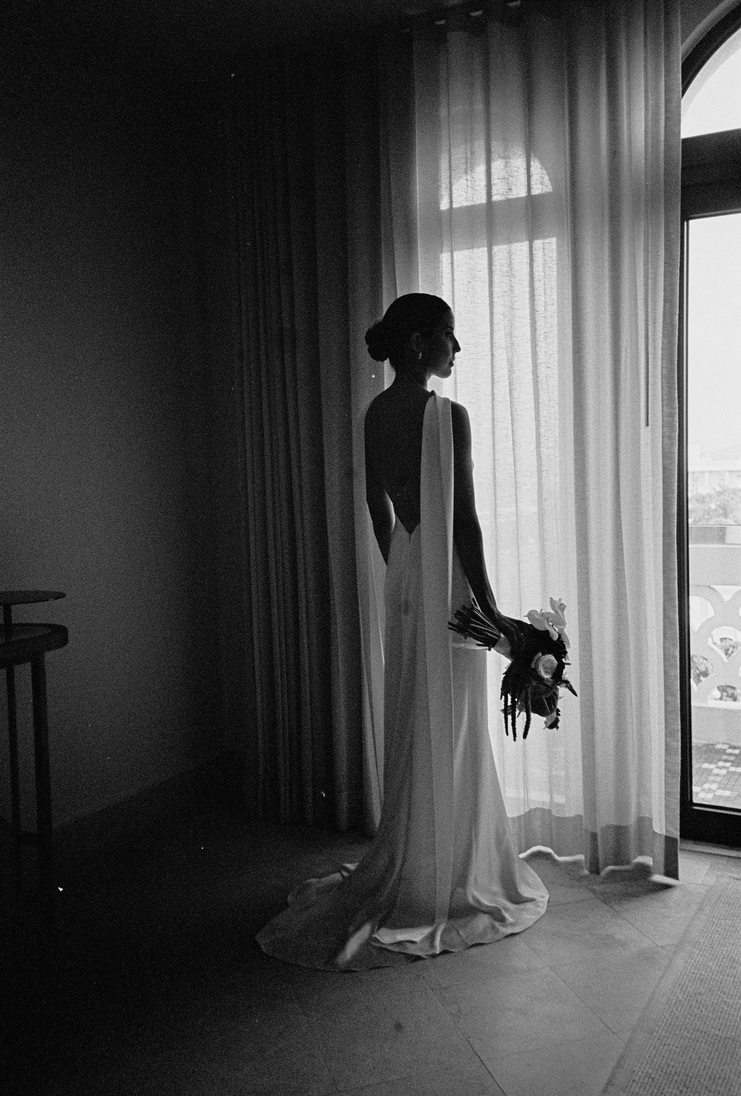 Elegant bride in backless white gown holding bouquet, standing by sheer curtains in black and white photo.