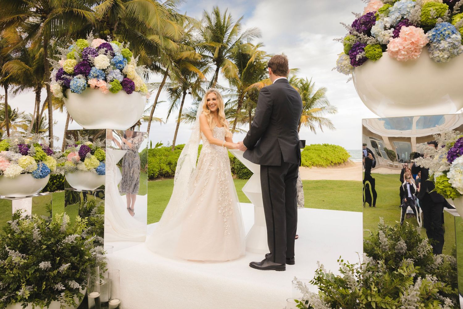 Wedding party gathers outdoors on a sunny day with palm trees and floral arrangements along a white path.