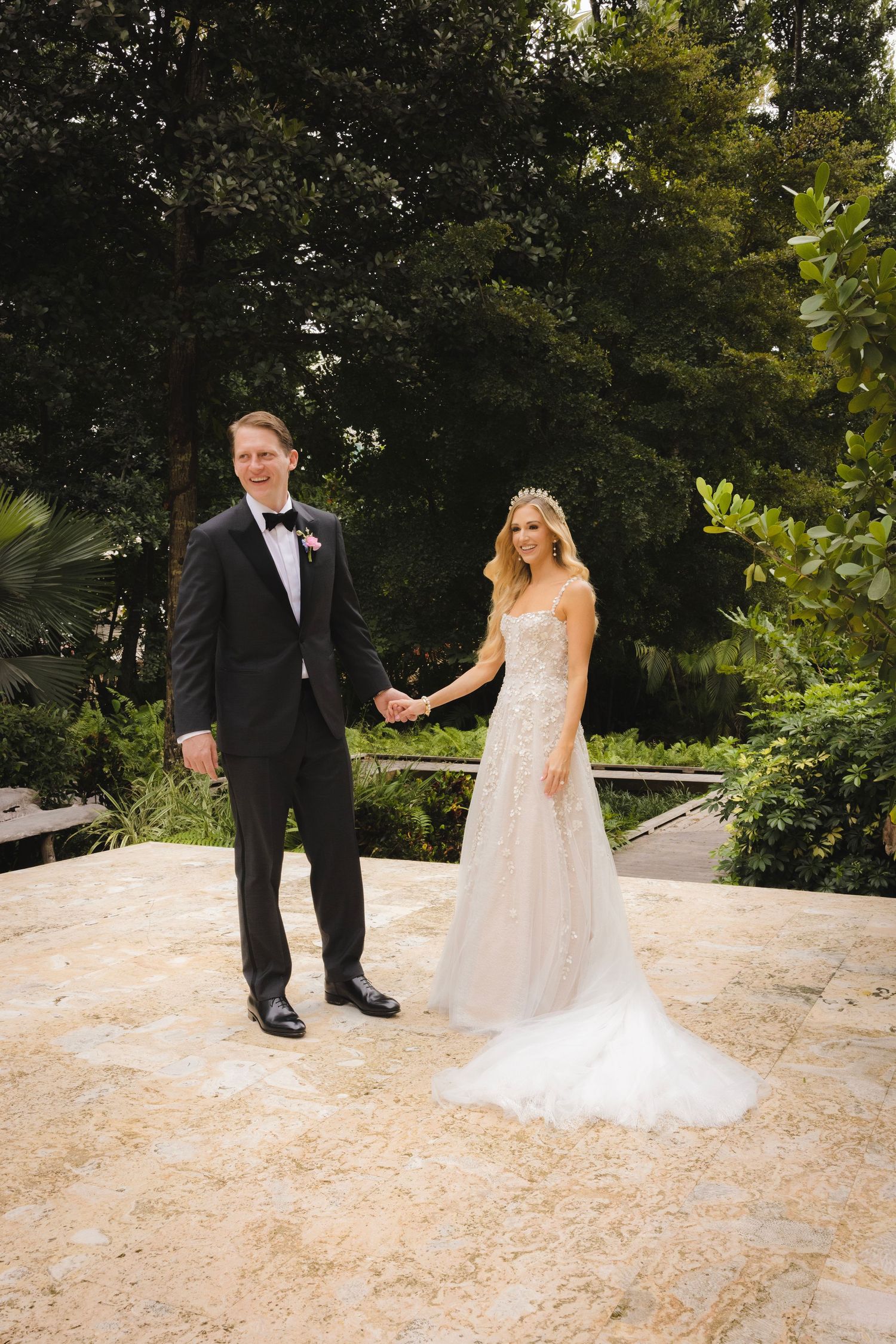 A couple in elegant wedding attire poses on a stone patio surrounded by tropical landscaping.