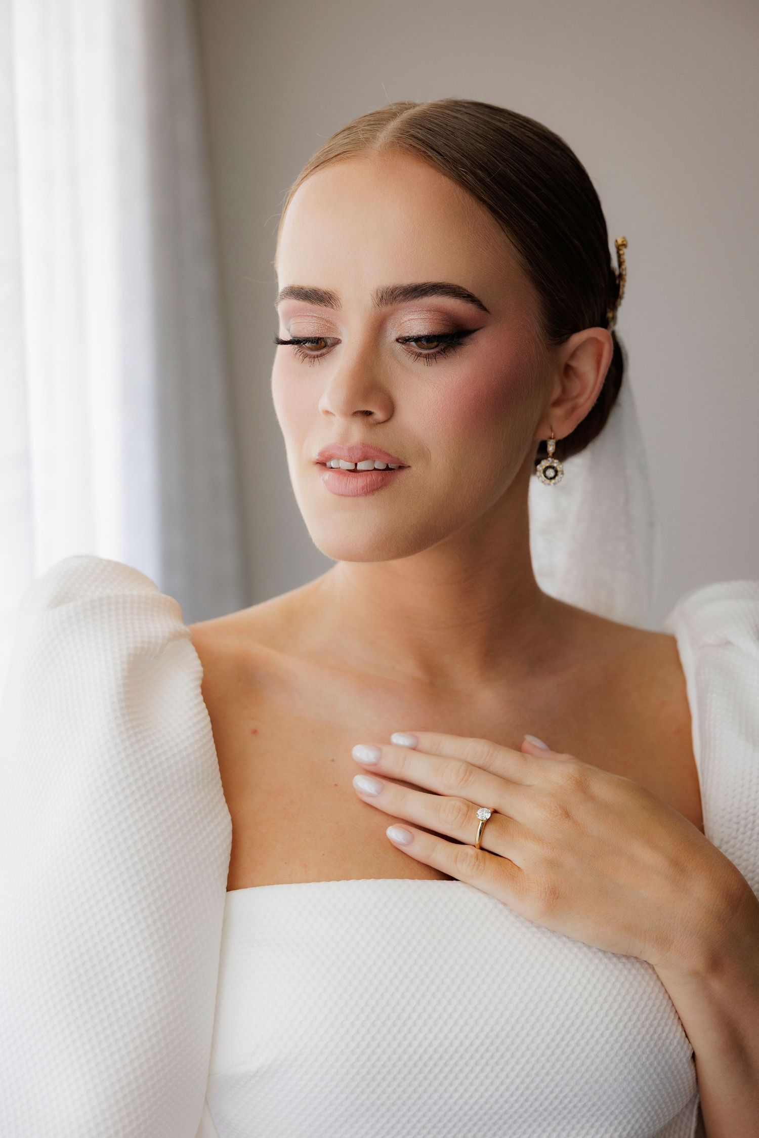 Beautiful bride in white gown with smokey eye makeup, sleek hairstyle and elegant jewelry shows off engagement ring.