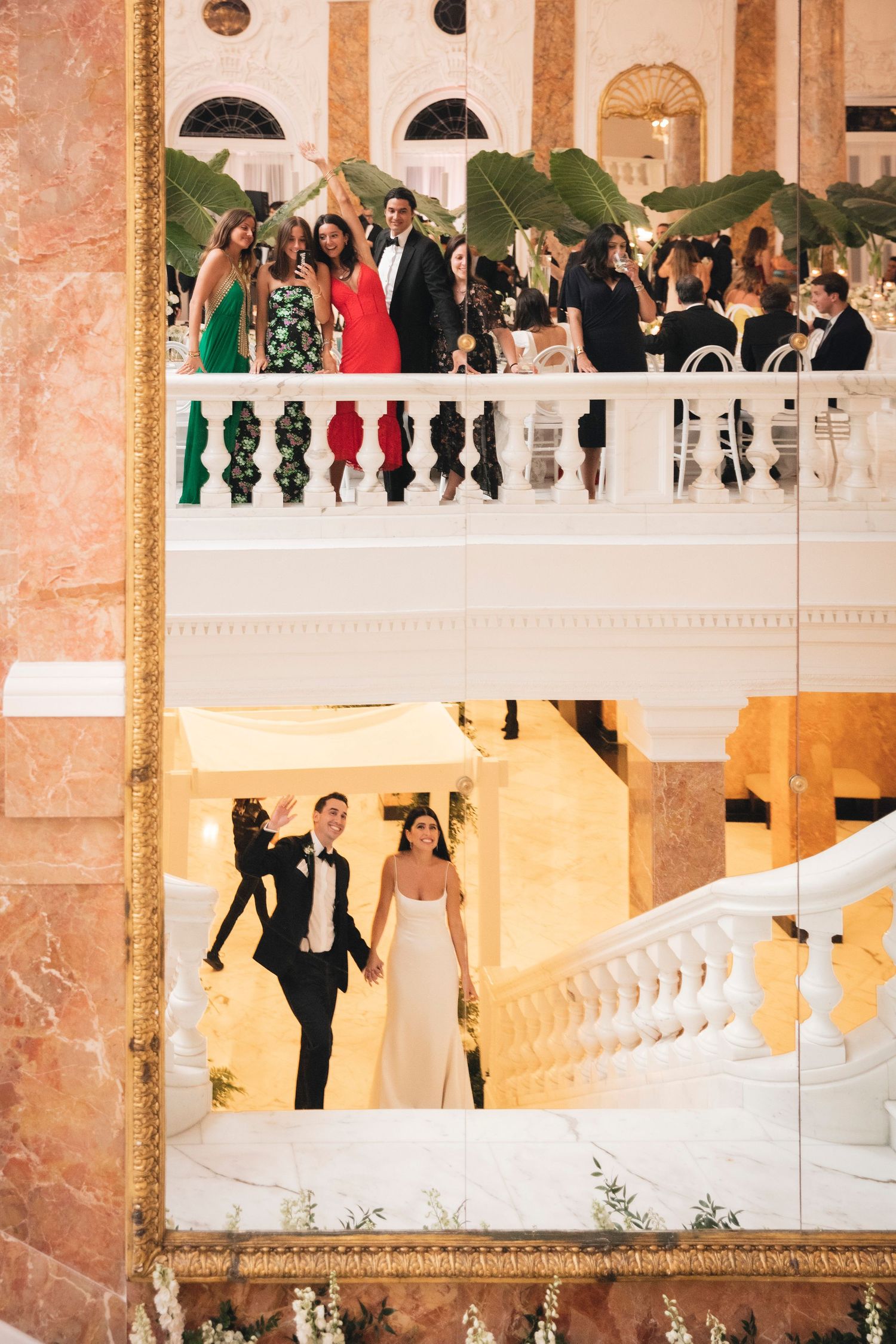 Wedding guests gather on an elegant marble staircase in a grand ballroom with coral-colored walls.