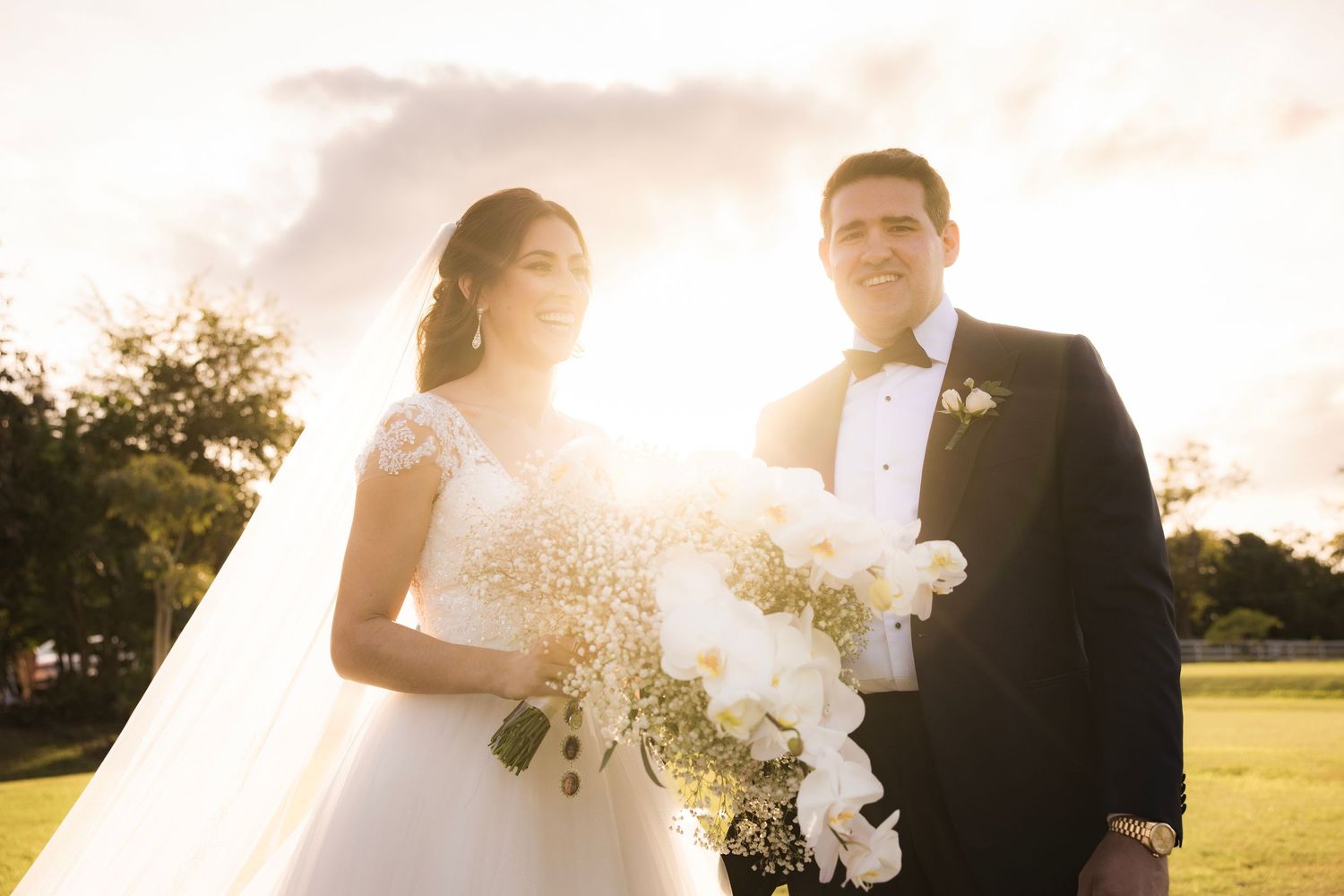 Romantic backlit wedding couple embraces in golden sunlight on a grassy field during sunset.