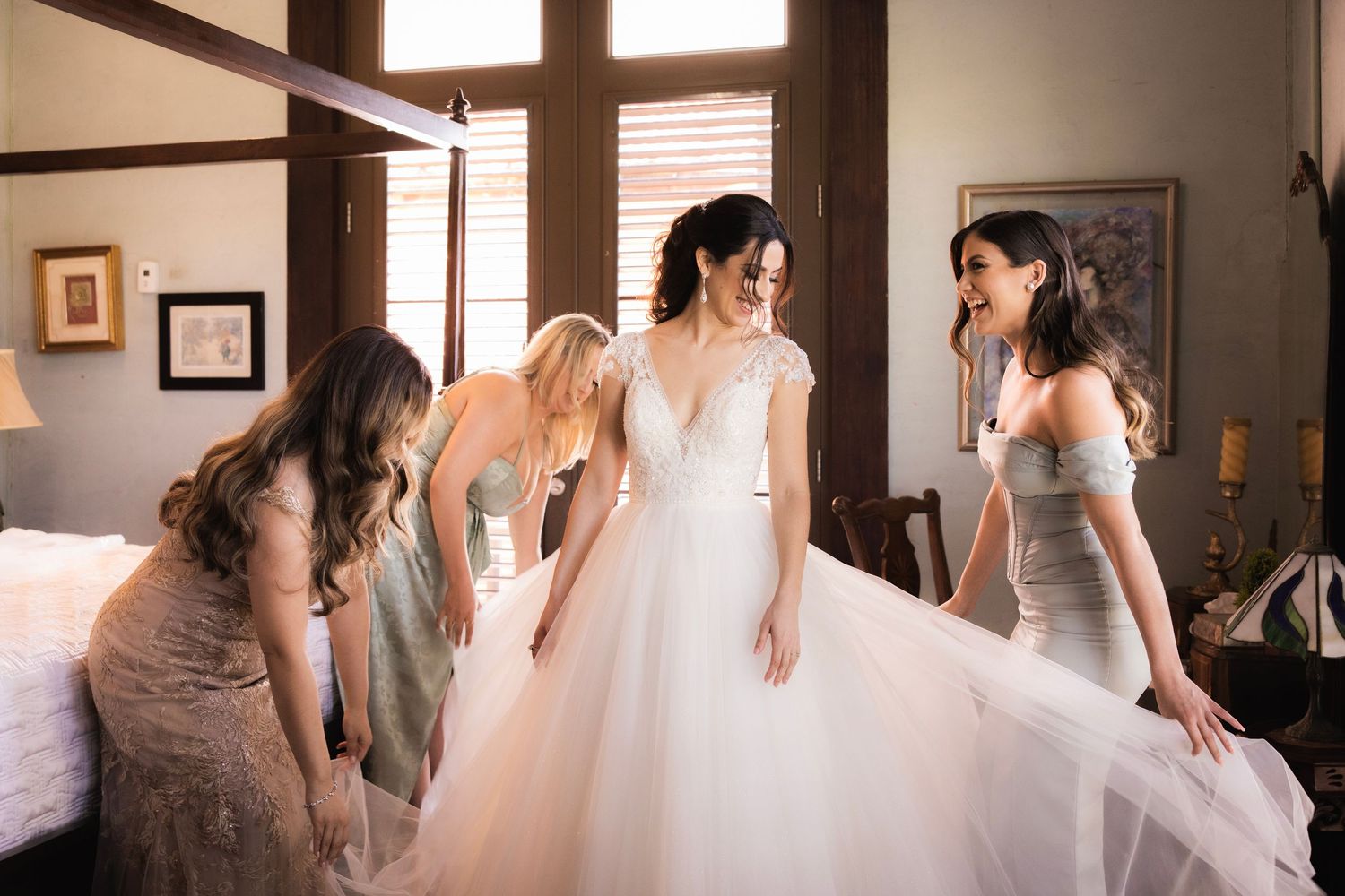 Bridesmaids help adjust the full tulle skirt of a white wedding gown in a sunlit bridal preparation room.