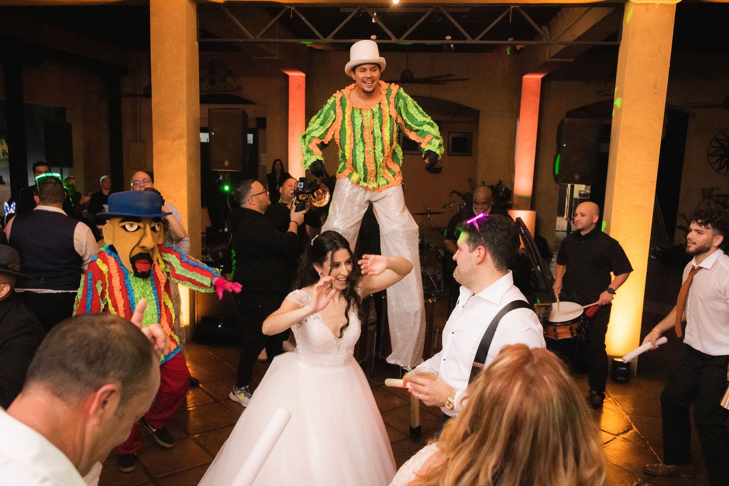 Wedding guests dance and celebrate on a dimly lit dance floor with people in formal attire and bright decorations.