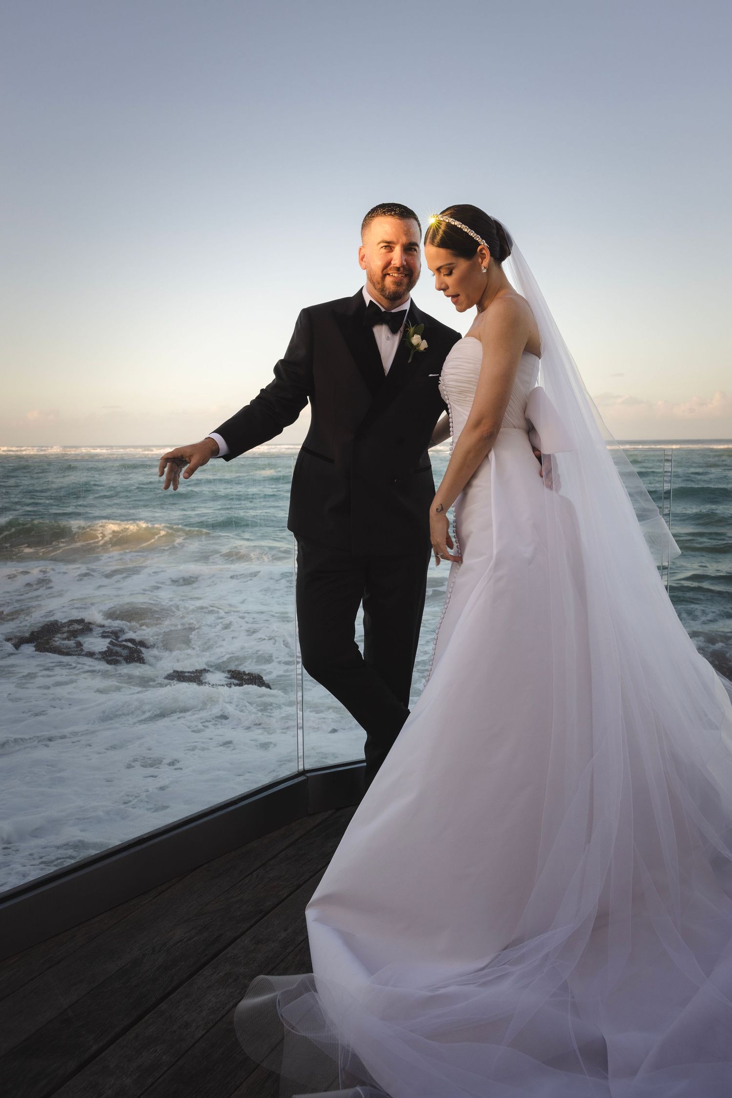A wedding couple stand together on a coastal pier at sunset with the ocean waves crashing behind them.