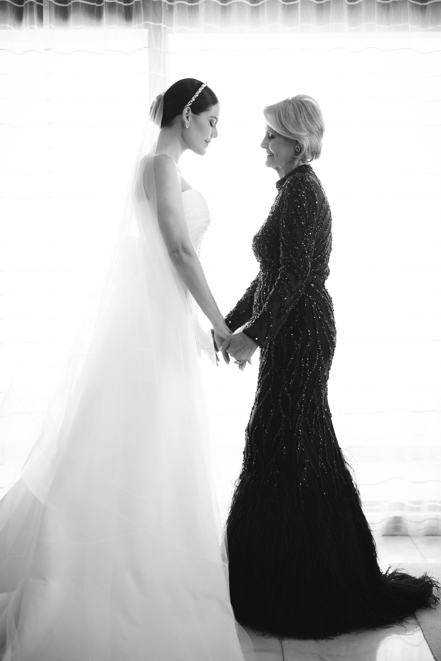 A touching black and white wedding moment shows a bride in white gown holding hands with woman in elegant black dress.