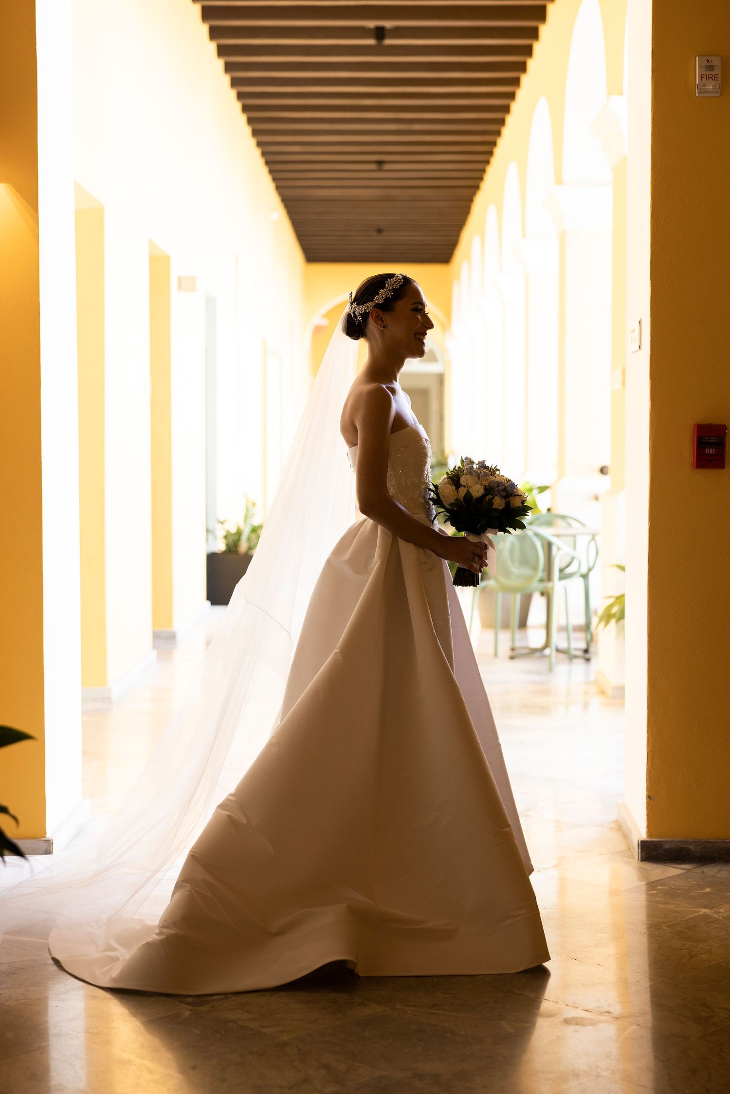 A bride in a flowing white gown poses elegantly in a colonnade hallway with warm lighting and pillars.