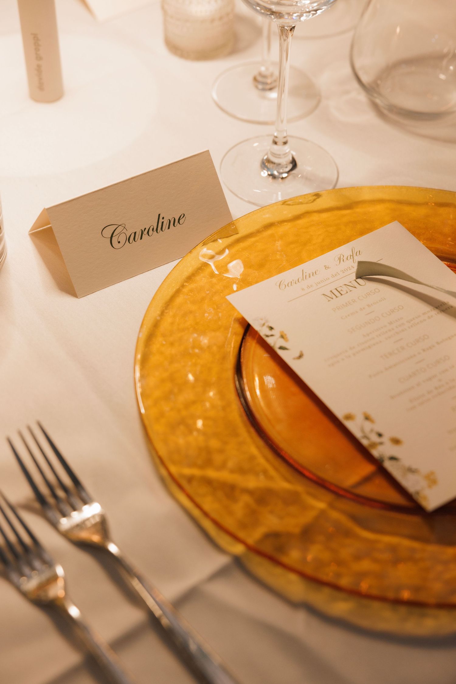 Elegant wedding place setting with amber glass charger plate, name card reading 'Caroline', and formal silverware on white linens.