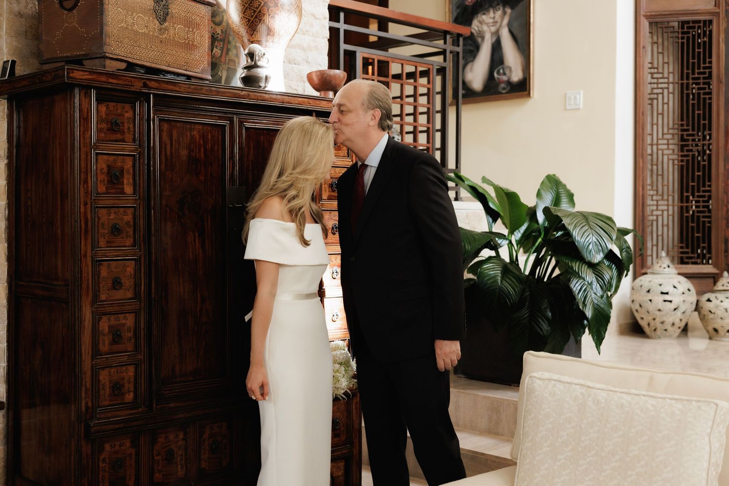 A romantic wedding moment at an indoor venue with elegant decor and a lush potted plant in the background.
