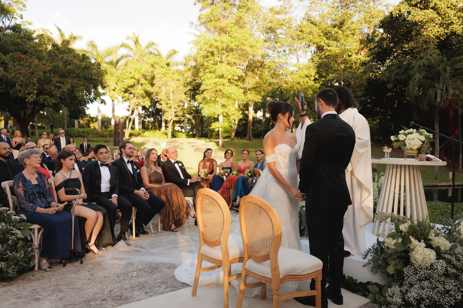 Wedding ceremony taking place outdoors under trees with guests seated on chairs and floral arrangements at sunset.