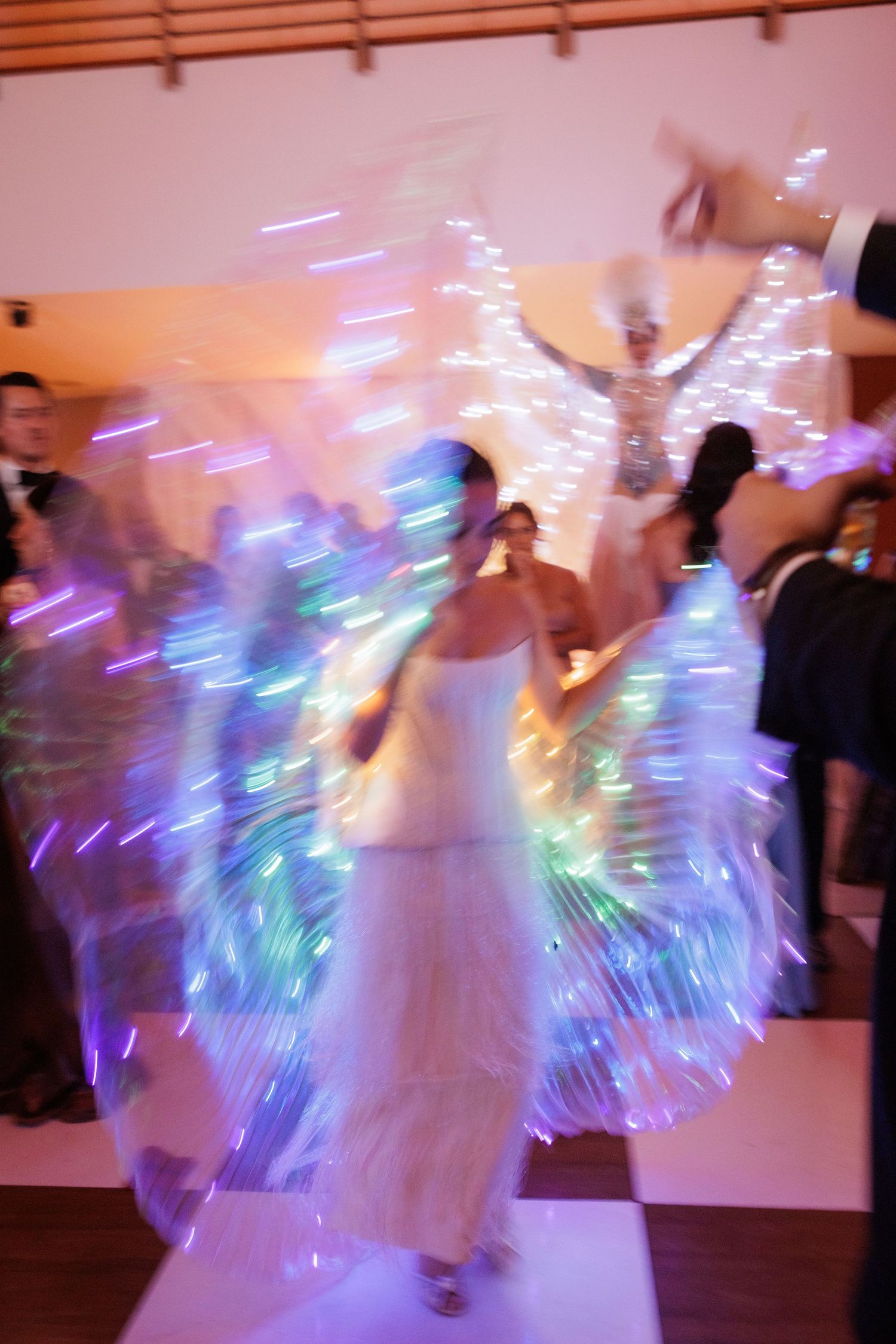 Motion-blurred photos show a dancer in white creating light trails with LED wings on a dance floor.