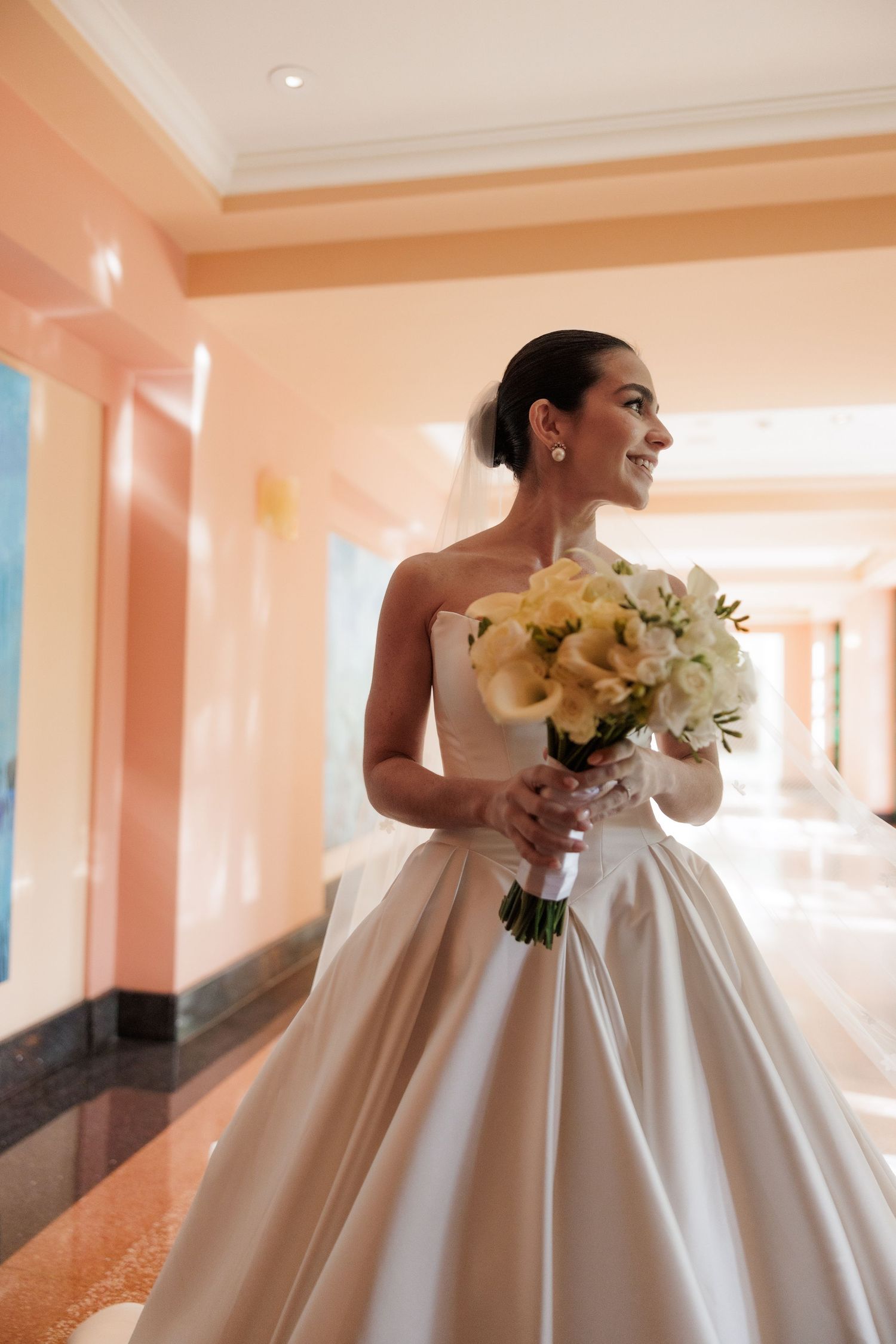 Bride in classic ballgown holds white bouquet in sunlit peach-colored hallway.