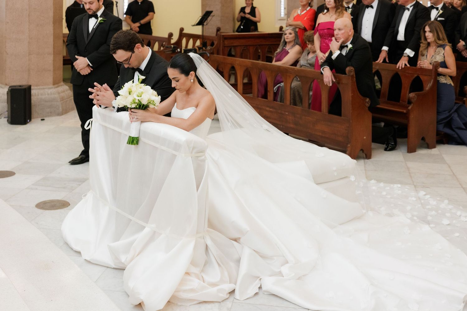 Wedding ceremony in a grand church with guests seated in wooden pews and white floral decorations along the aisle.