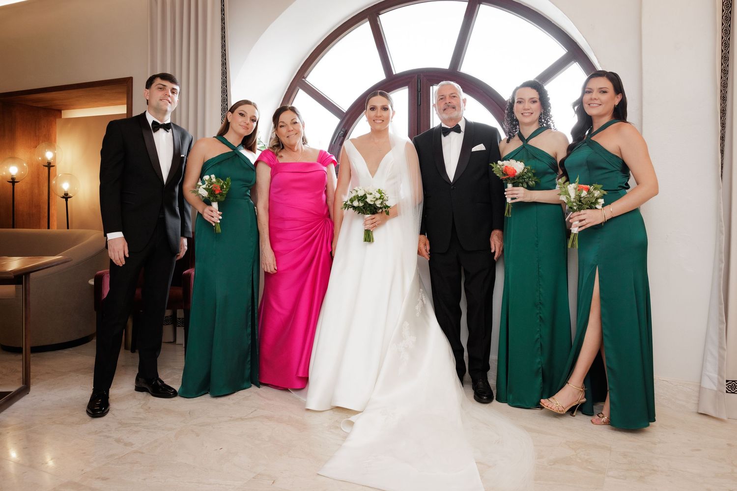 Wedding party poses elegantly in front of arched windows wearing emerald green and pink formal attire.