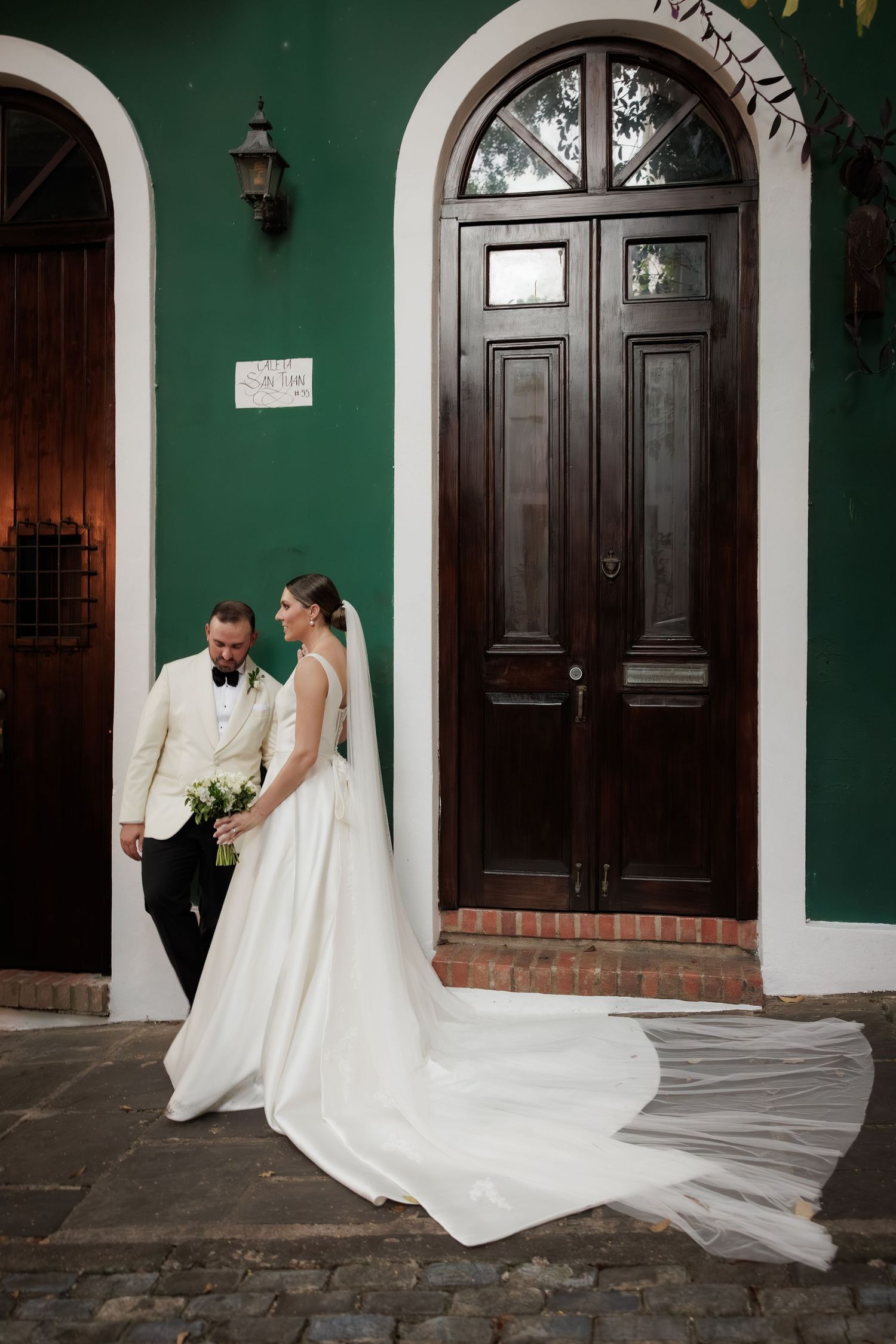 A wedding couple stands by an ornate green colonial doorway with the bride's long white train flowing behind them.